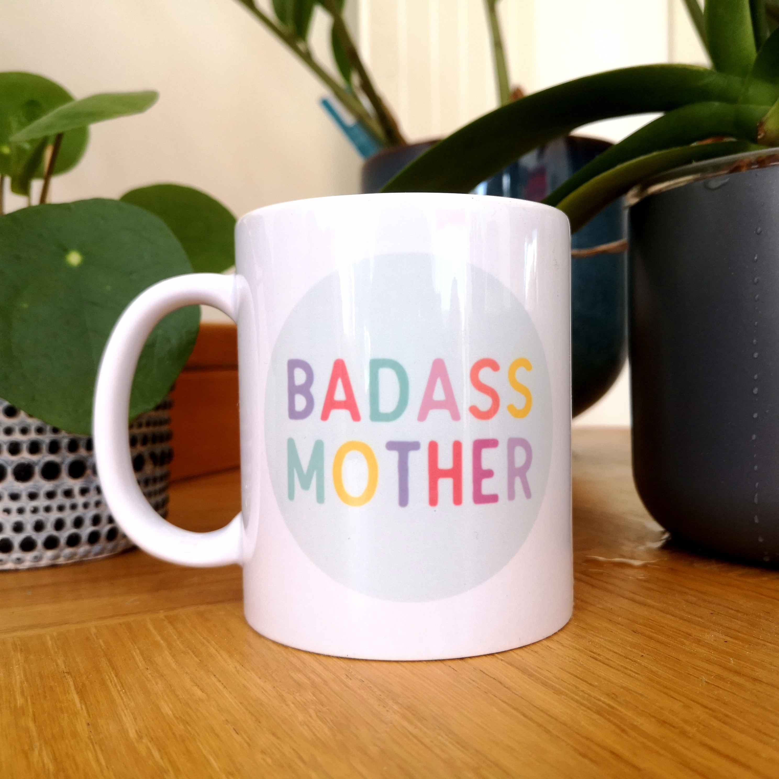 A white 10oz ceramic mug with BADASS MOTHER in bright rainbow colours on a pale blue oval