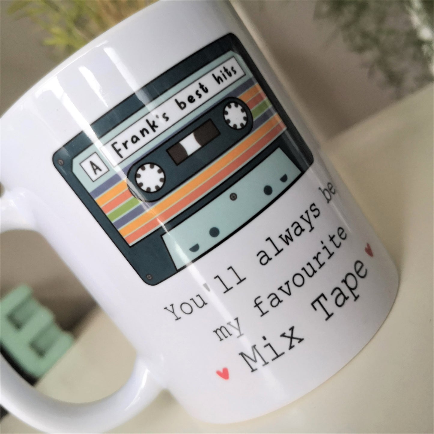 A close up of a  white ceramic mug with a picture of an old cassette tape with a personalised message written on it, then underneath the tap is 'You'll always be my favourite mix tape' with 2 red hearts either side of it