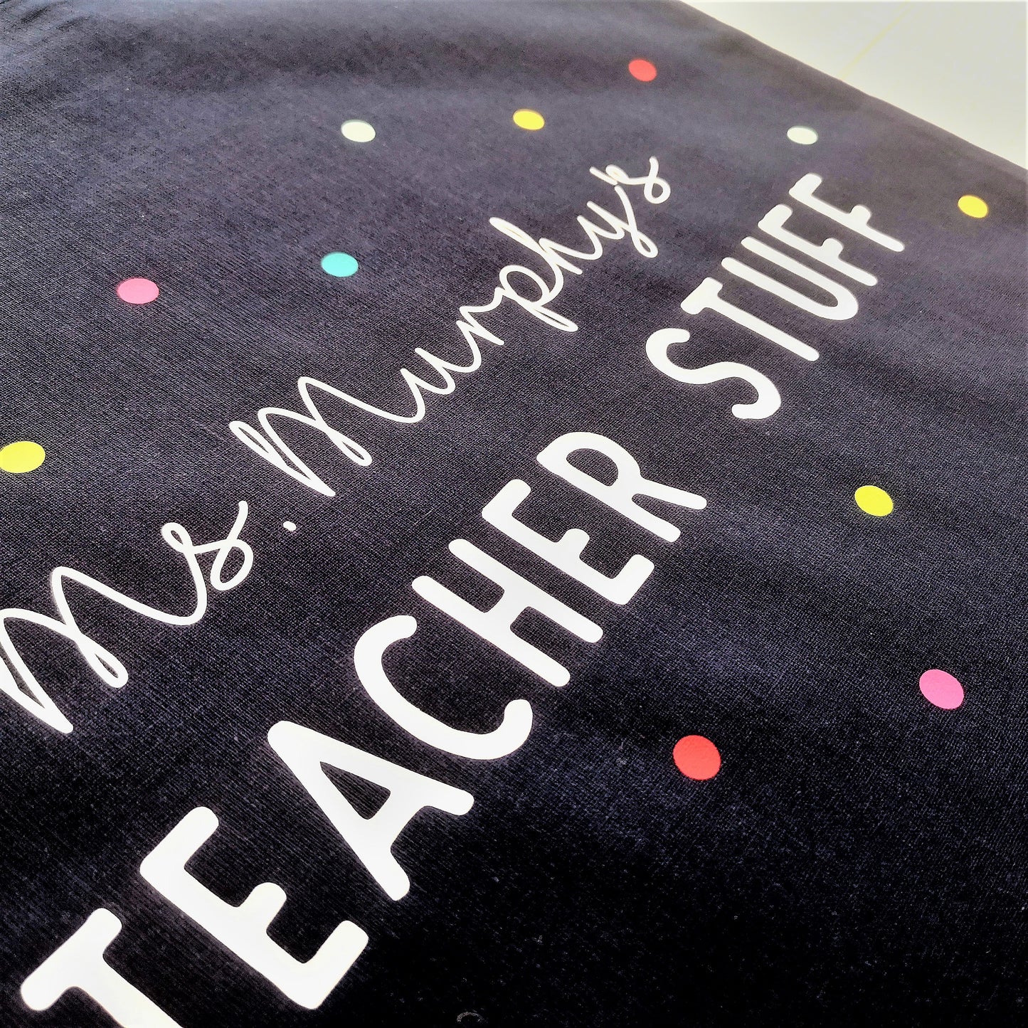 A close up photo of a personalised lightweight black cotton tote with <Name>'s TEACHER STUFF on it. The text is surrounded with multicoloured polkadots