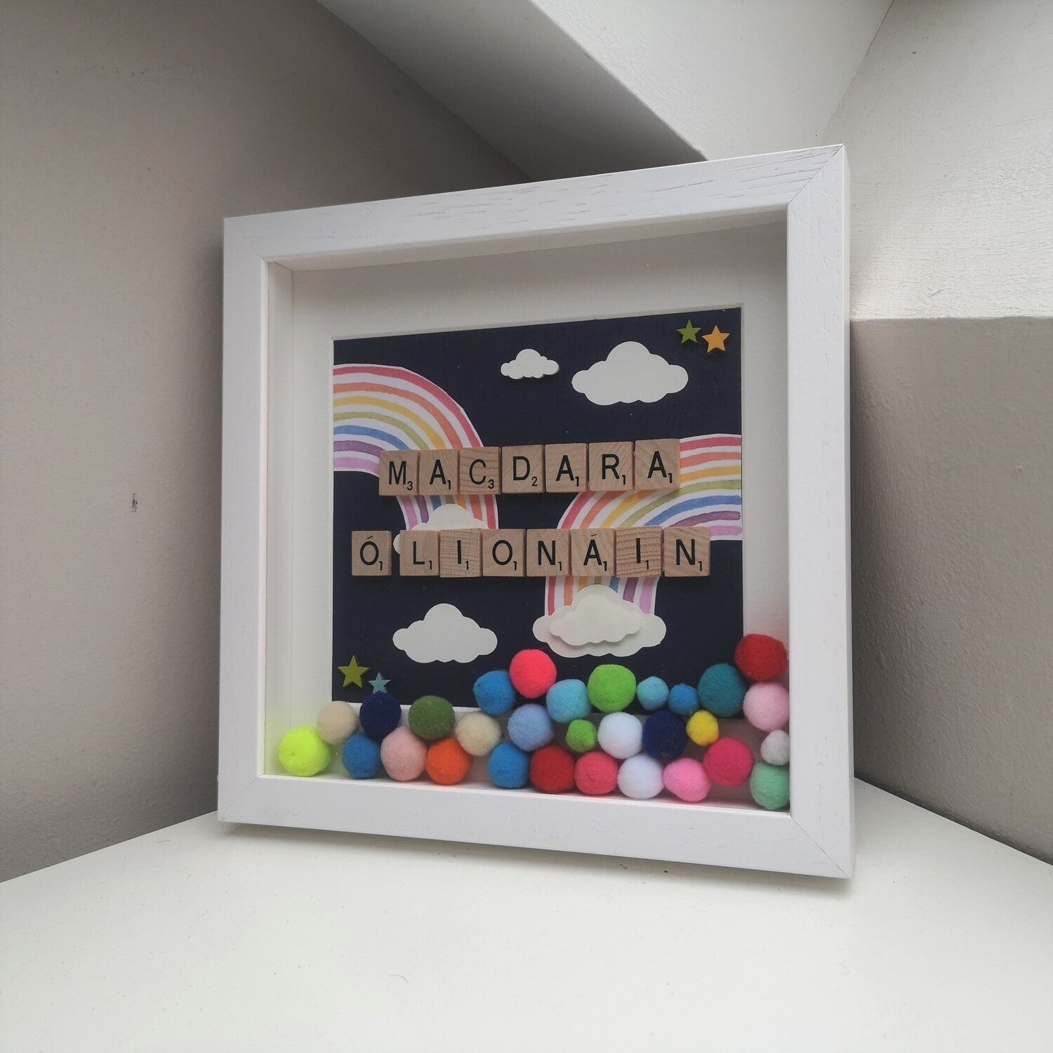 A name in wooden tiles with Papercut Rainbows and clouds, on a navy background, bright coloured pompoms and wooden clouds in a handmade 25x25cm deep box wooden frame.
