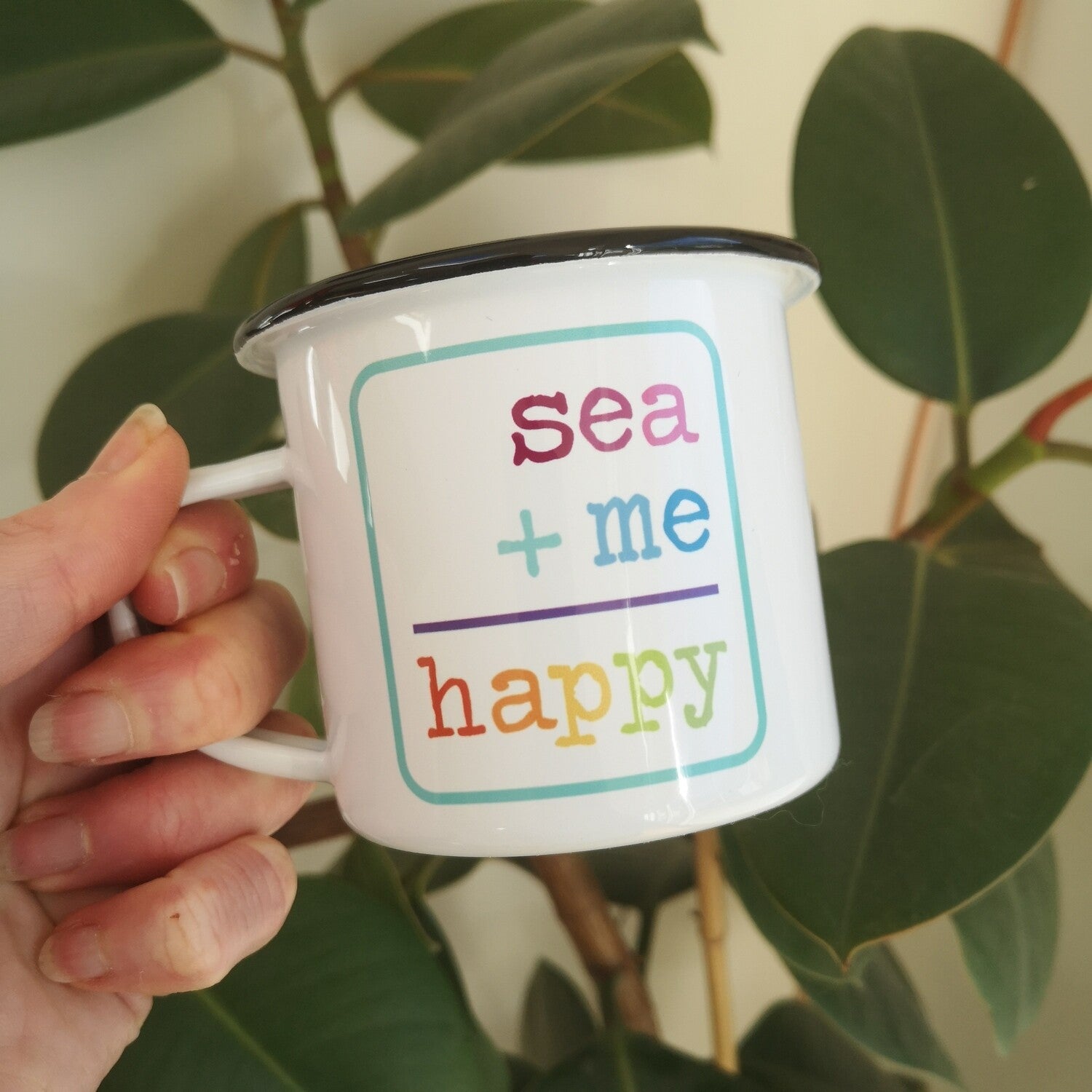 Another view of a White enamel mug with a black rim with the following on the front - SEA + ME = HAPPY in rainbow colours