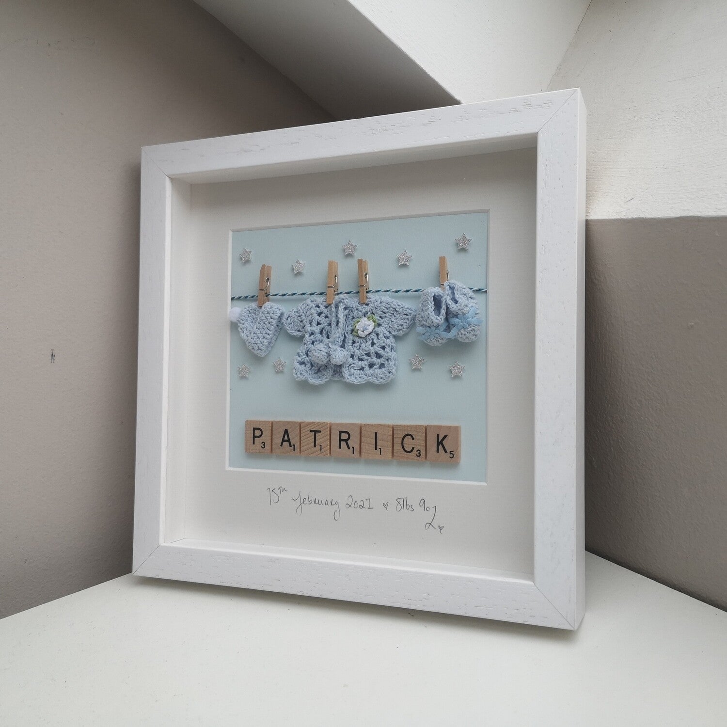 A personalised baby blue washing line with crochet hat, cardie & booties, with a name in wooden scrabble tiles below the washing line, in a handmade 25x25cm deep box wooden frame.