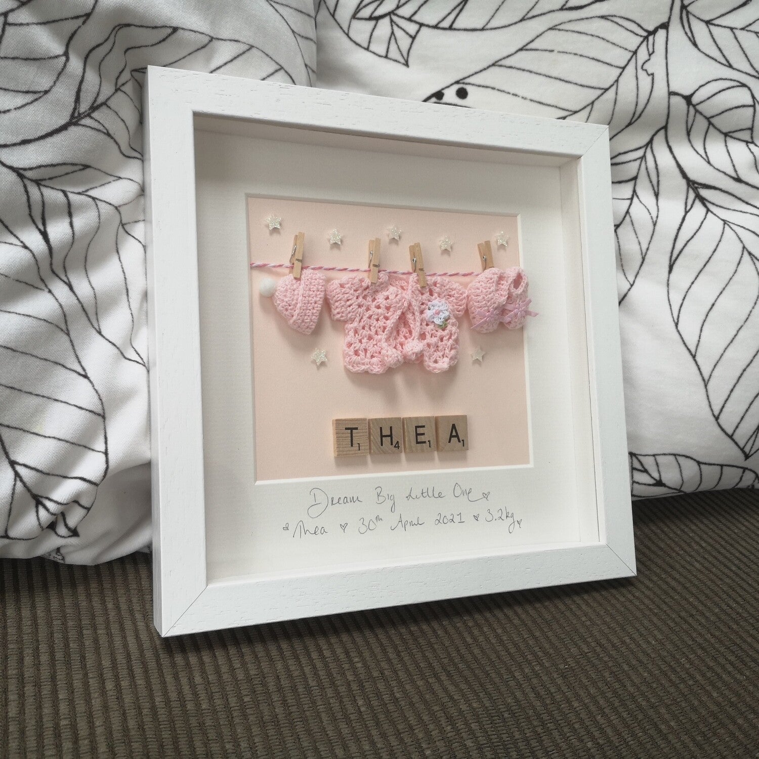 A personalised baby pink washing line with crochet hat, cardie & booties, with a name in wooden scrabble tiles below the washing line, in a handmade 25x25cm deep box wooden frame.