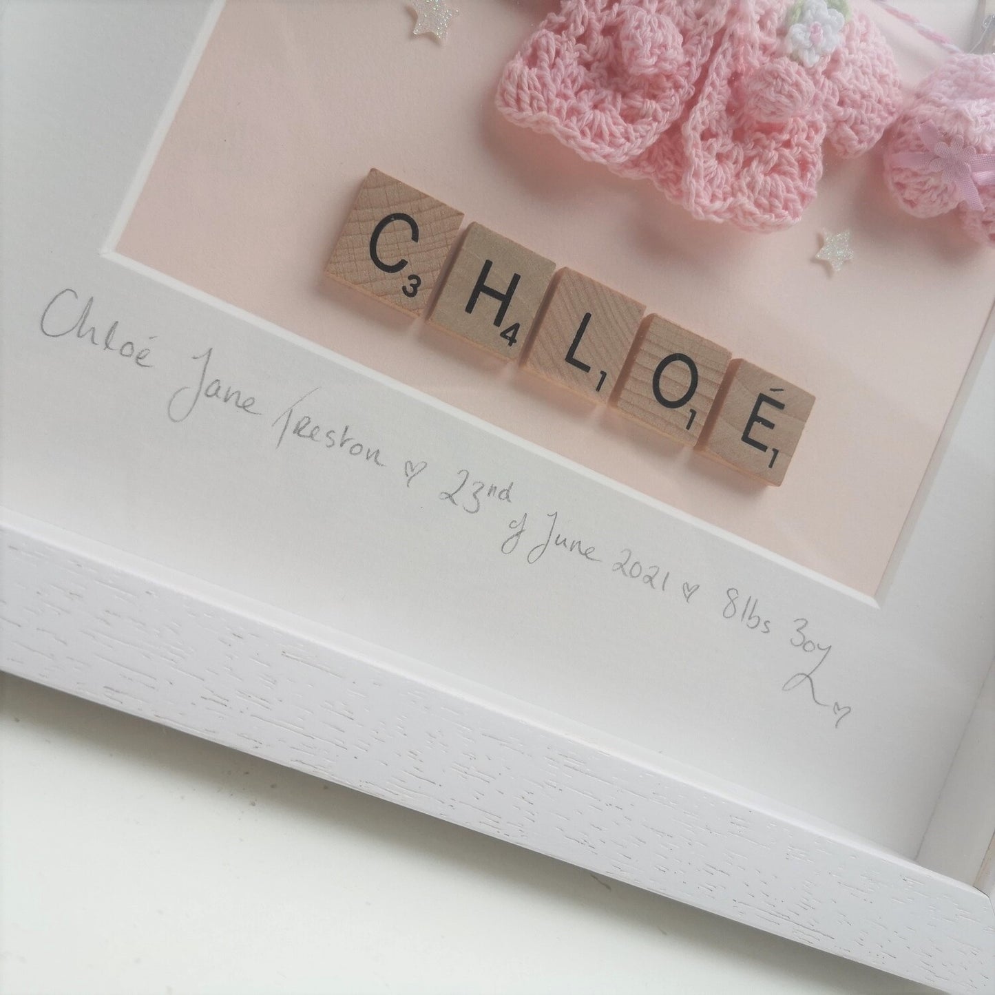 A close up of A personalised baby pink washing line with crochet hat, cardie & booties, with a name in wooden scrabble tiles below the washing line, in a handmade 25x25cm deep box wooden frame.