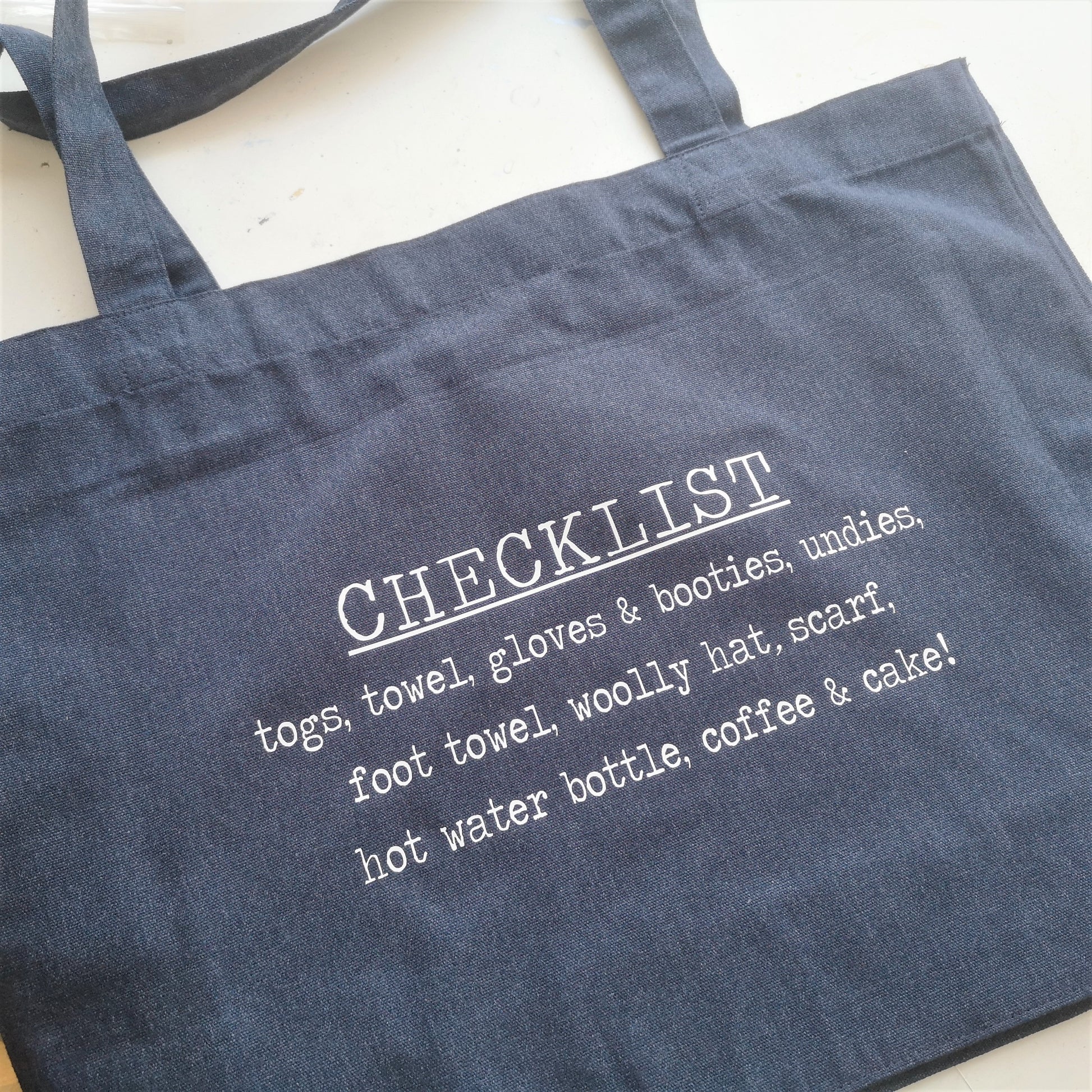 A close up of a midnight blue organic cotton tote with toggle closing with the following text on the front in white type font - a personalised checklist so you won't forget to pack anything for swimming! 