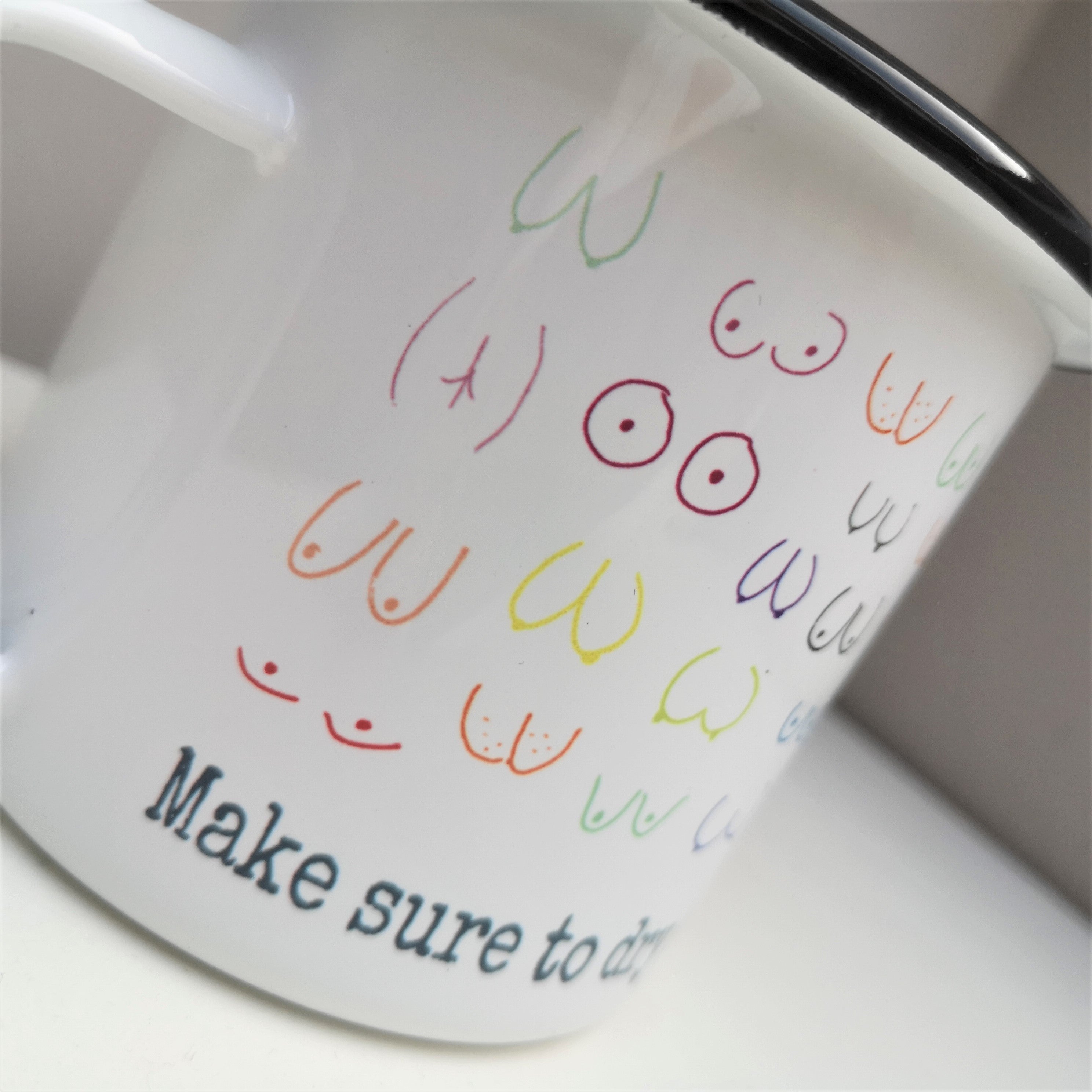 A close up of a white enamel mug with a black rim with handdrawn boobs and bums on it and the following slogan below the drawings - Make sure to dry all your wobbly bits and don't forget your pants