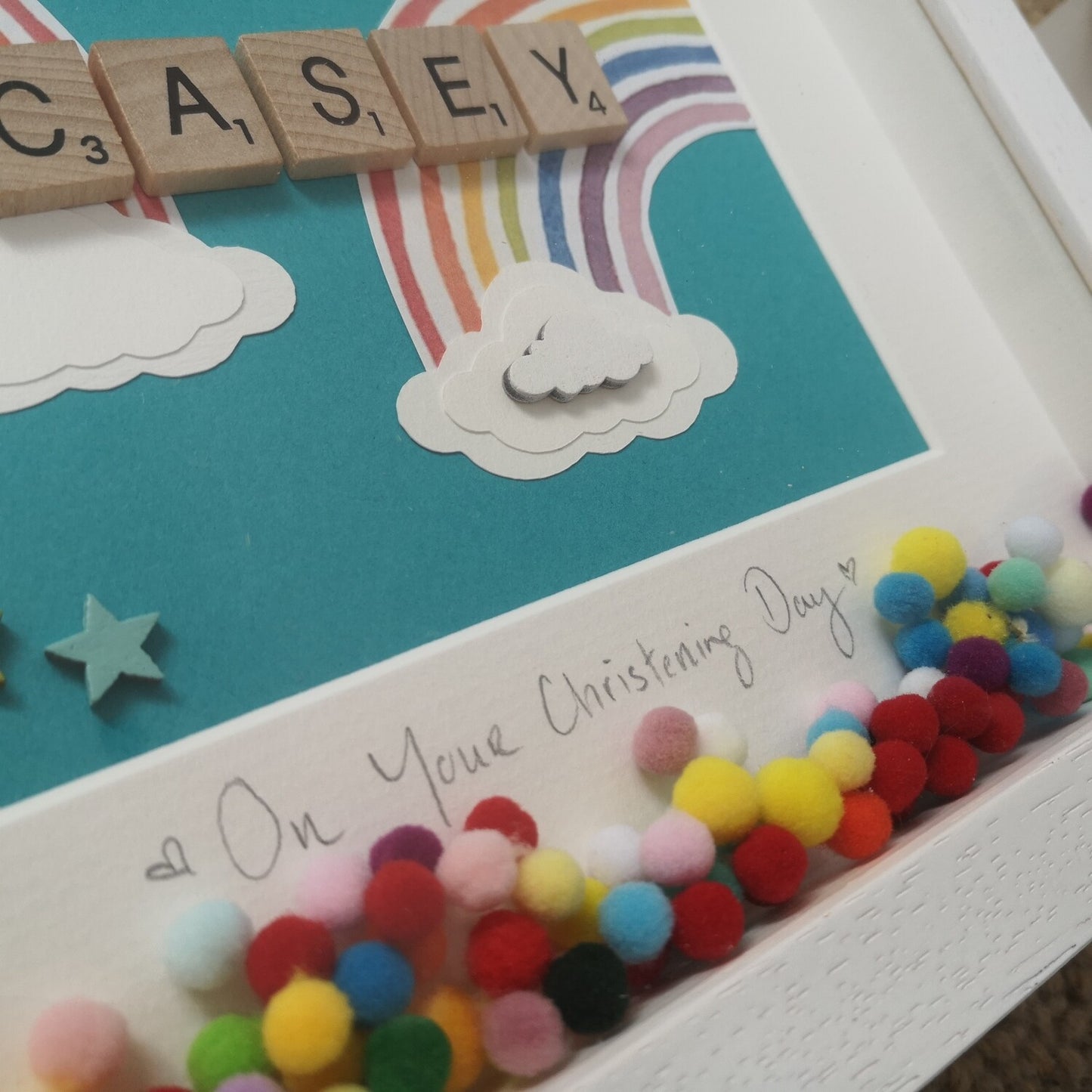 A close up photo of a name in wooden tiles on a  teal background with Papercut Rainbows and clouds, bright coloured pompoms and wooden clouds in a handmade 25x25cm deep box wooden frame.
