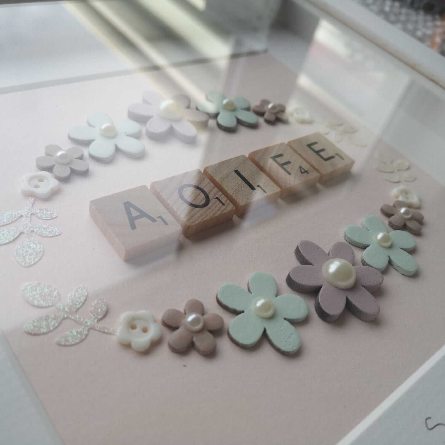 A close up of a pretty pastel wreath frame with a name in the middle in wooden letter tiles. The wreath is made with wooden flowers, pearls resin buttons and sparkly card leaves, in a handmade 25x25cm deep box wooden frame.
