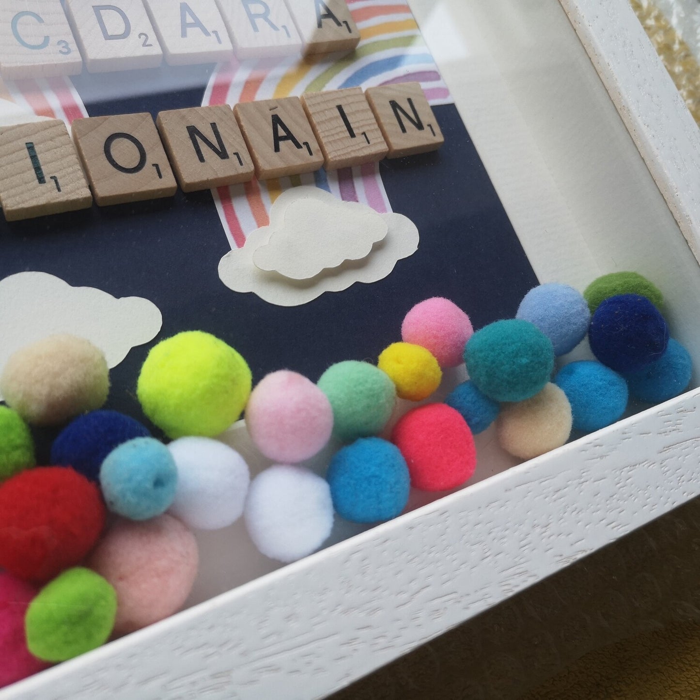 A close up of the name in wooden tiles with Papercut Rainbows and clouds, on a navy background, bright coloured pompoms and wooden clouds in a handmade 25x25cm deep box wooden frame.
