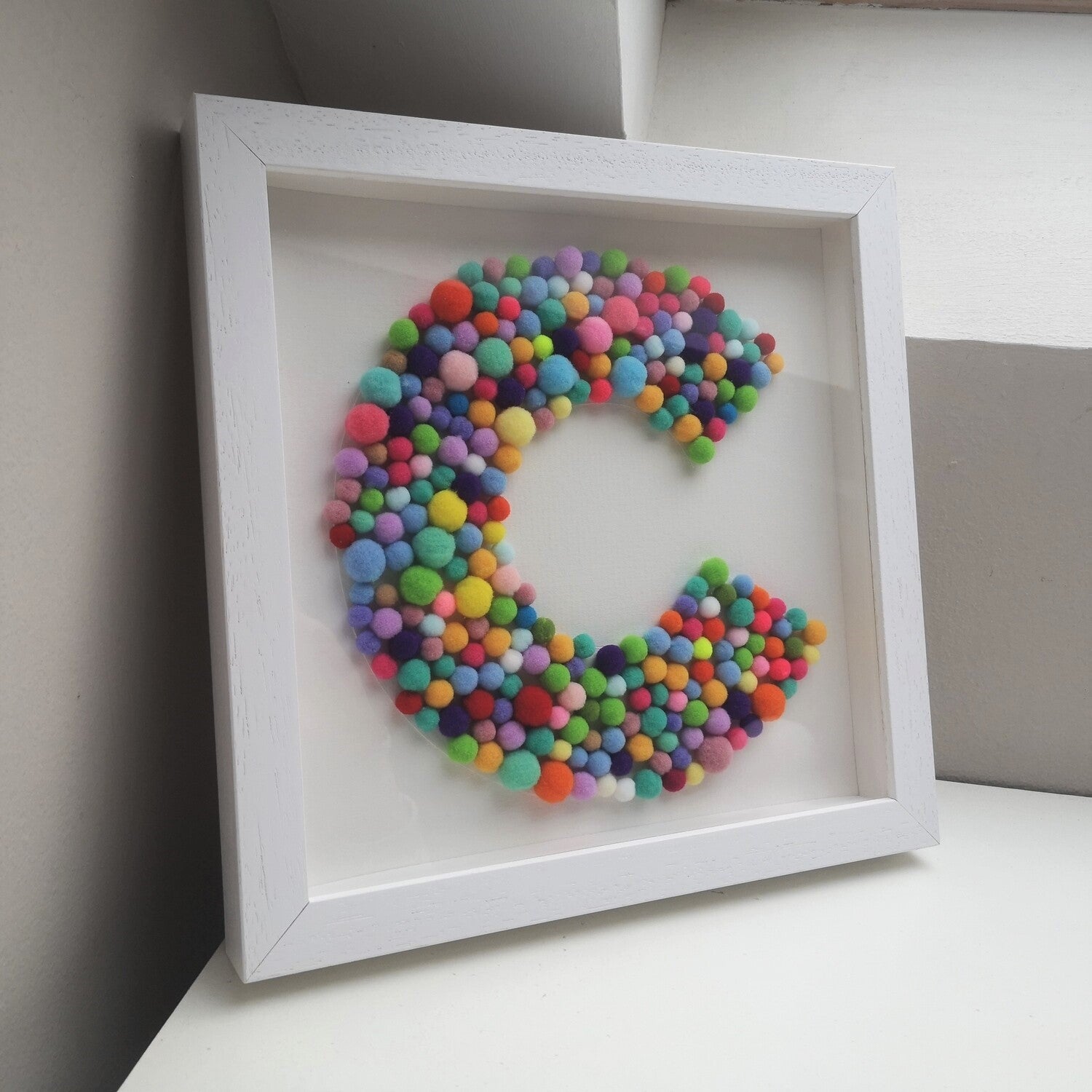 A colourful tactile rainbow pompom Initial frame, in a handmade wooden deep box frame