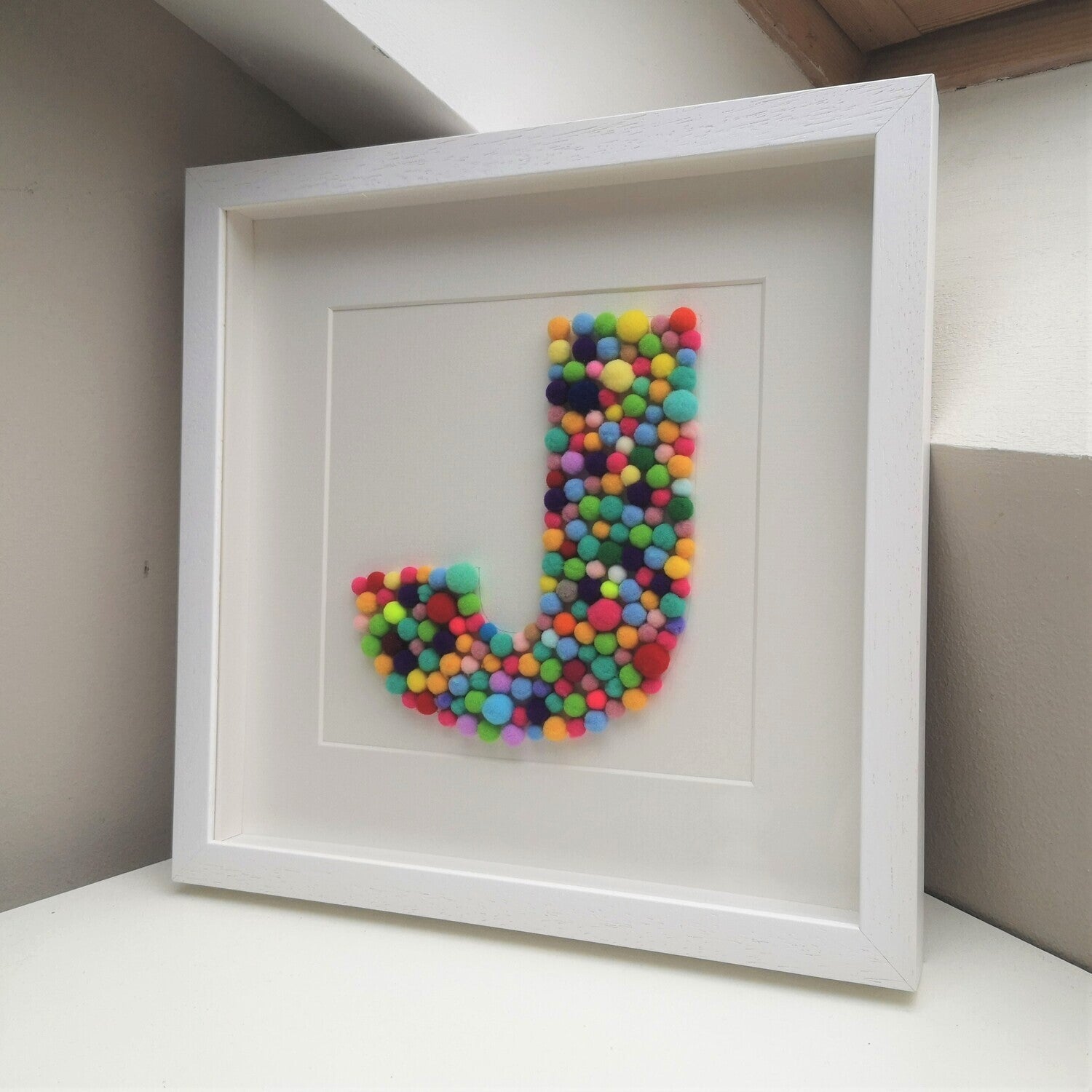 A colourful tactile rainbow pompom Initial frame, in the larger 30x30cm handmade wooden deep box frame