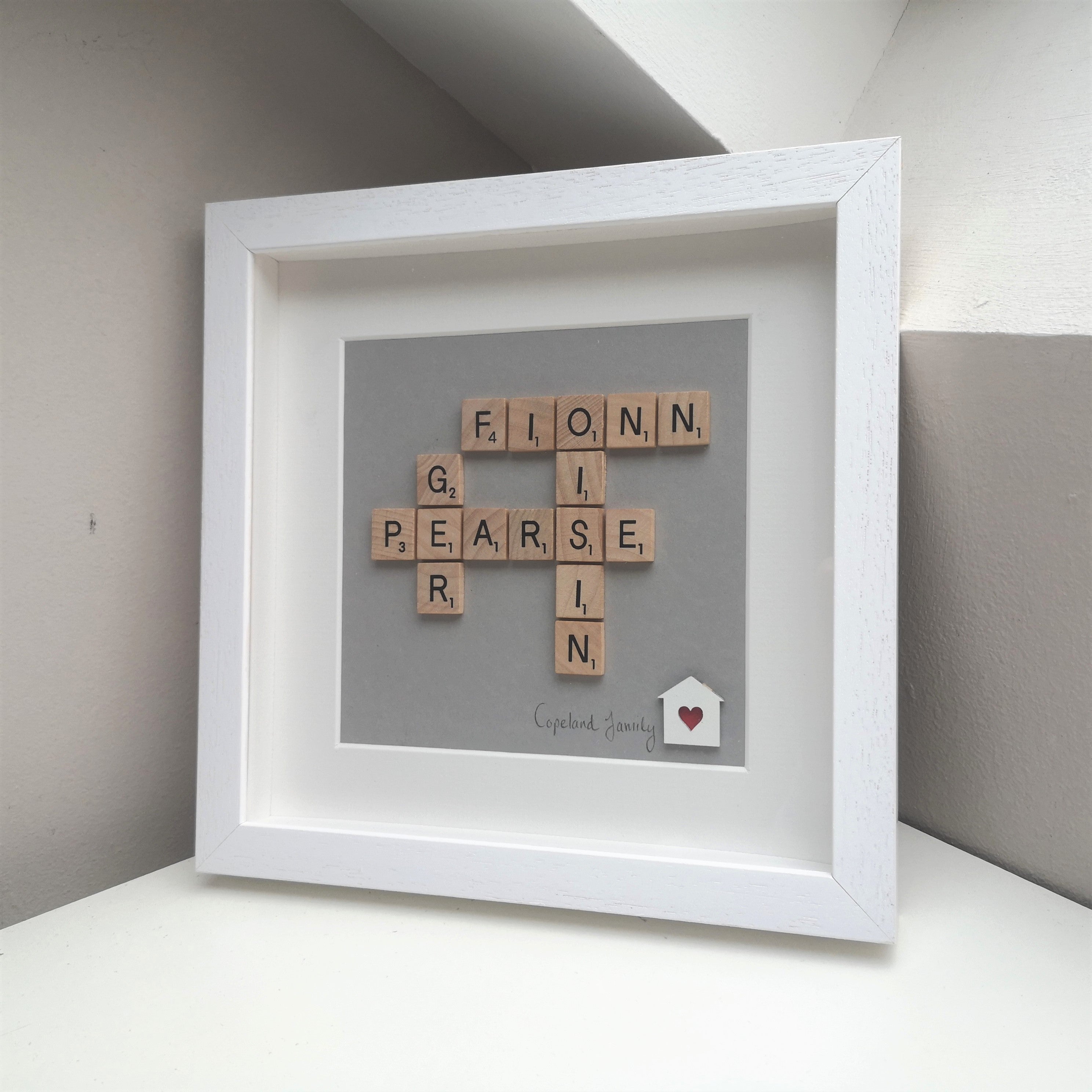 Family Names in wooden scrabble tiles on grey background with little wooden house and heart heartcut in a handmade white 25x25cm deep box wooden frame