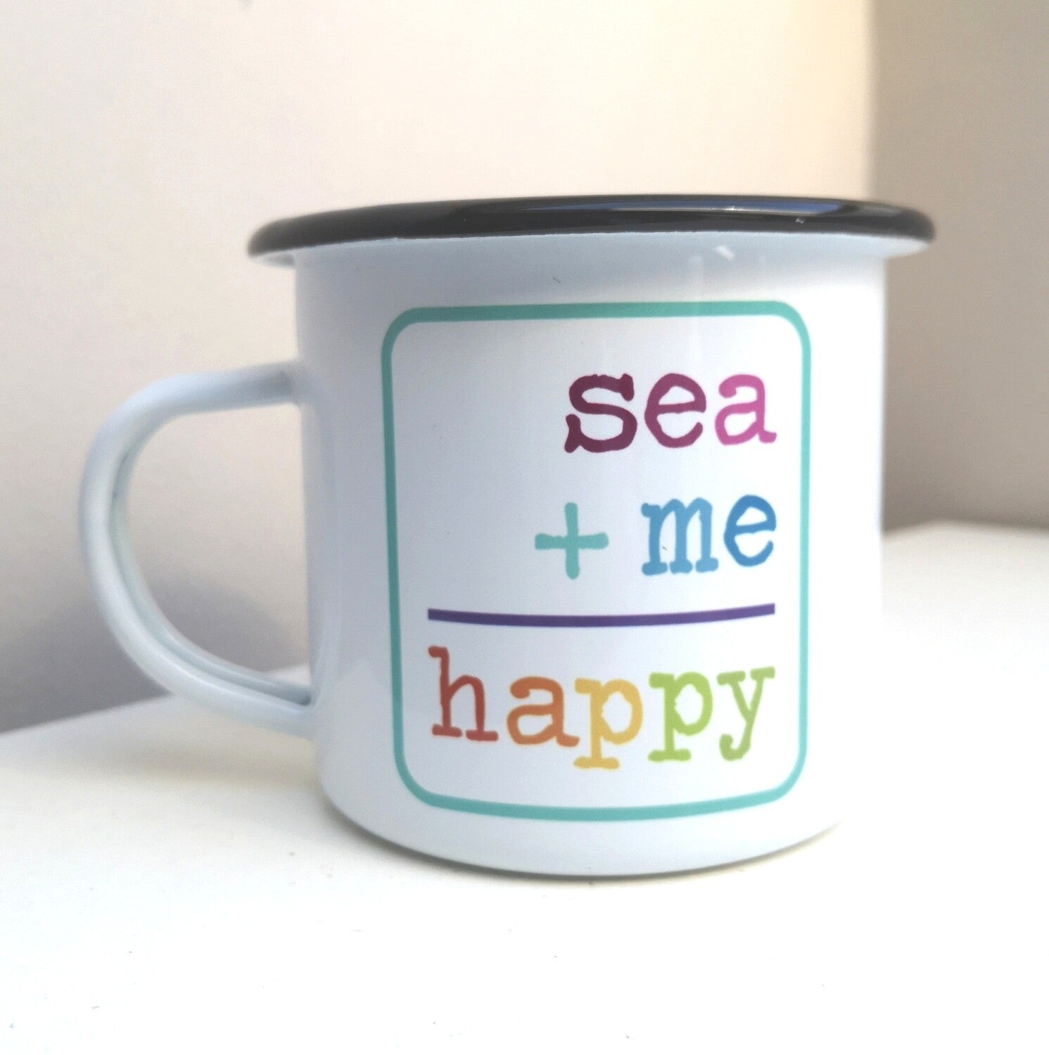 A White enamel mug with a black rim with the following on the front - SEA + ME = HAPPY in rainbow colours