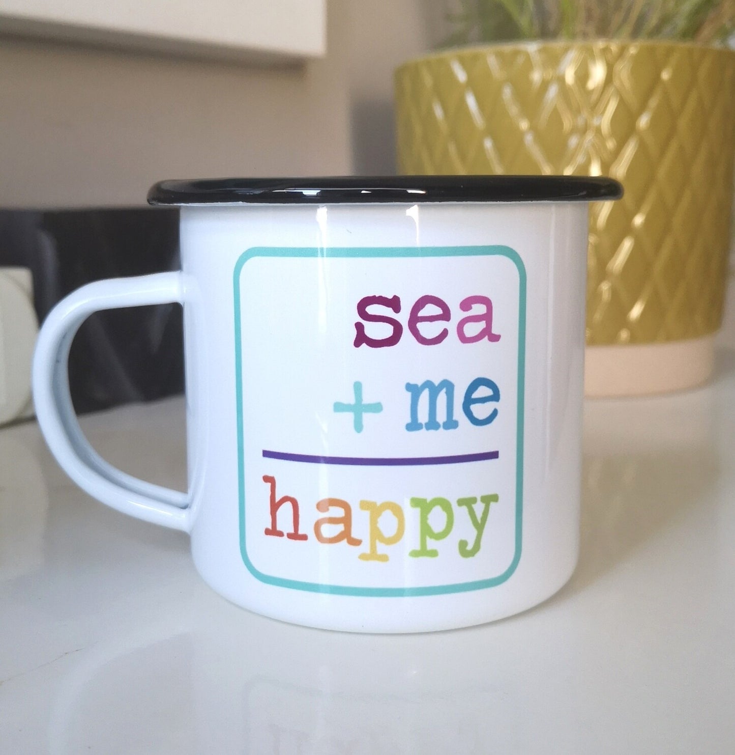 A White enamel mug with a black rim in front of a plant pot, with the following on the front - SEA + ME = HAPPY in rainbow colours