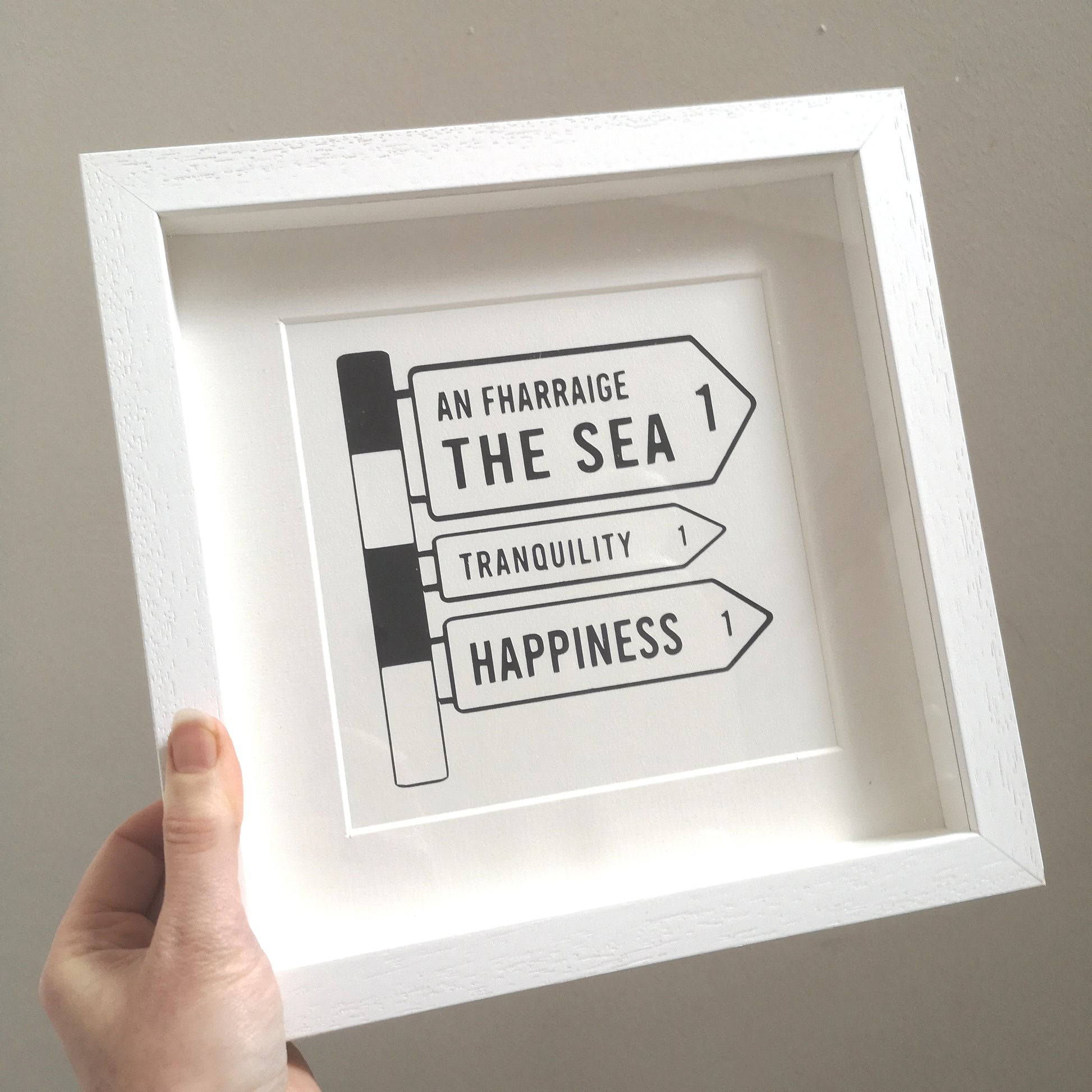 A hand holding up a 25x25cm deep box white wooden frame with a papercutting in it of an old Irish Roadsign pointing to the right with arrows for The Sea, Tranquility and Happiness