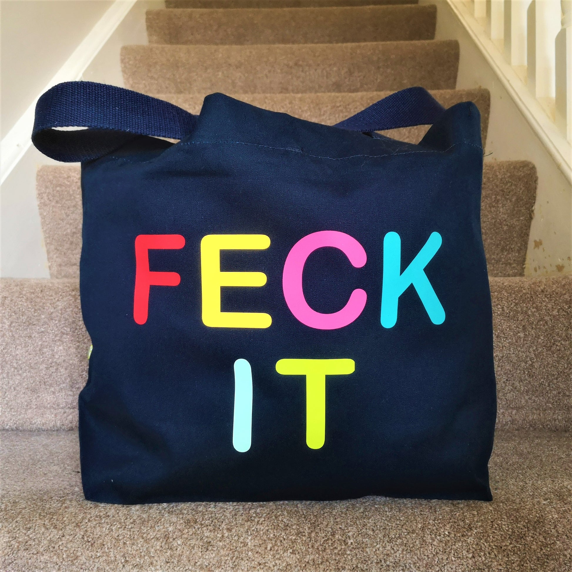 A navy organic cotton tote with toggle closing with the following text on the front in rainbow coloured type font - FECK IT 
