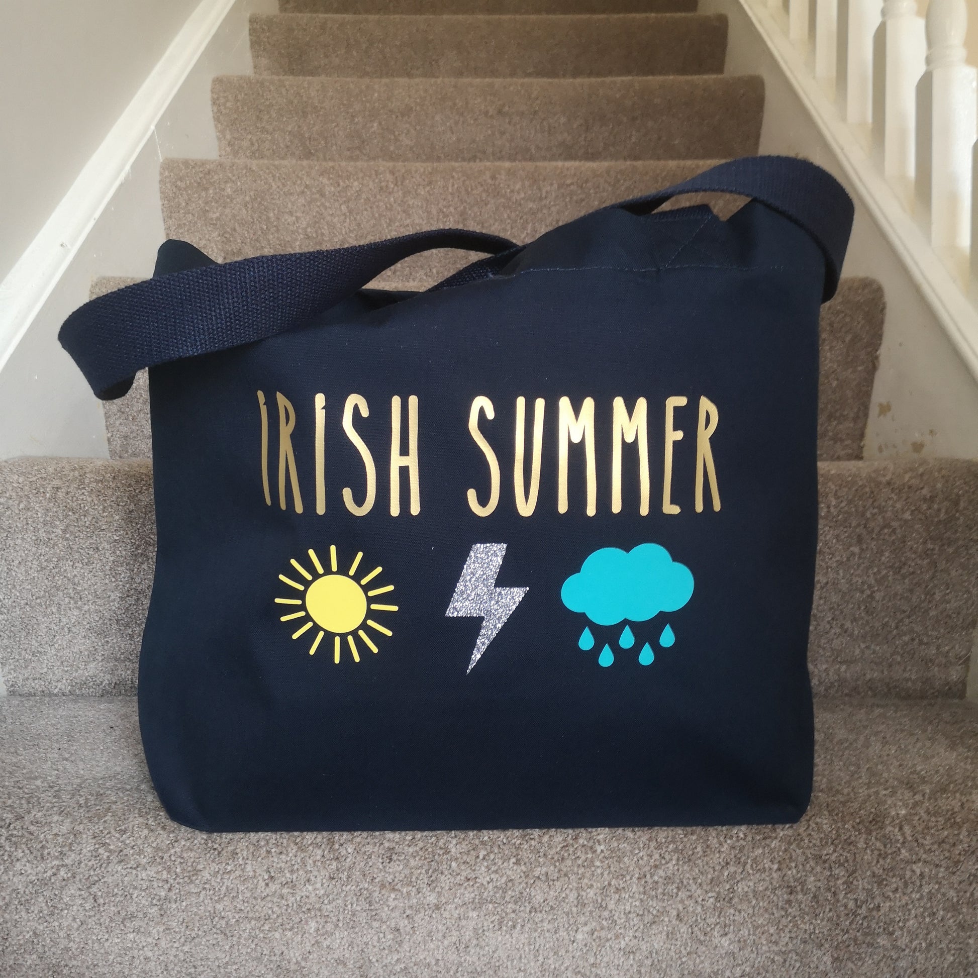 A black organic cotton tote with toggle closing with the following text on the front in gold font - IRISH SUMMER. Below the text is a yellow sun, a silver lightening bolt and a light blue rain cloud 