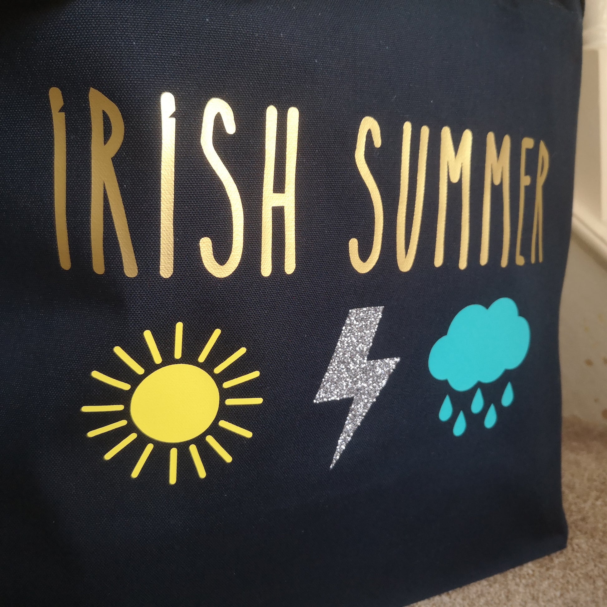 A close up of a black organic cotton tote with toggle closing with the following text on the front in gold font - IRISH SUMMER. Below the text is a yellow sun, a silver lightening bolt and a light blue rain cloud 
