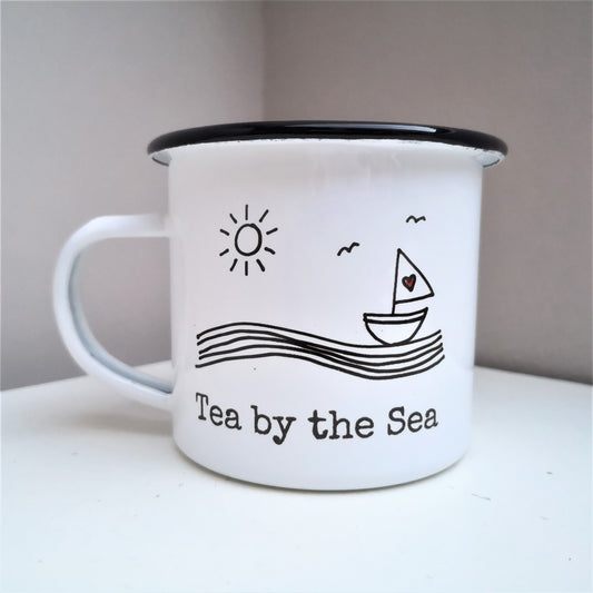 A White enamel mug with a black rim with the following on the front - a hand drawn seaside scene with sailboat, waves, bird and sun, with the following text below it - Tea by the Sea