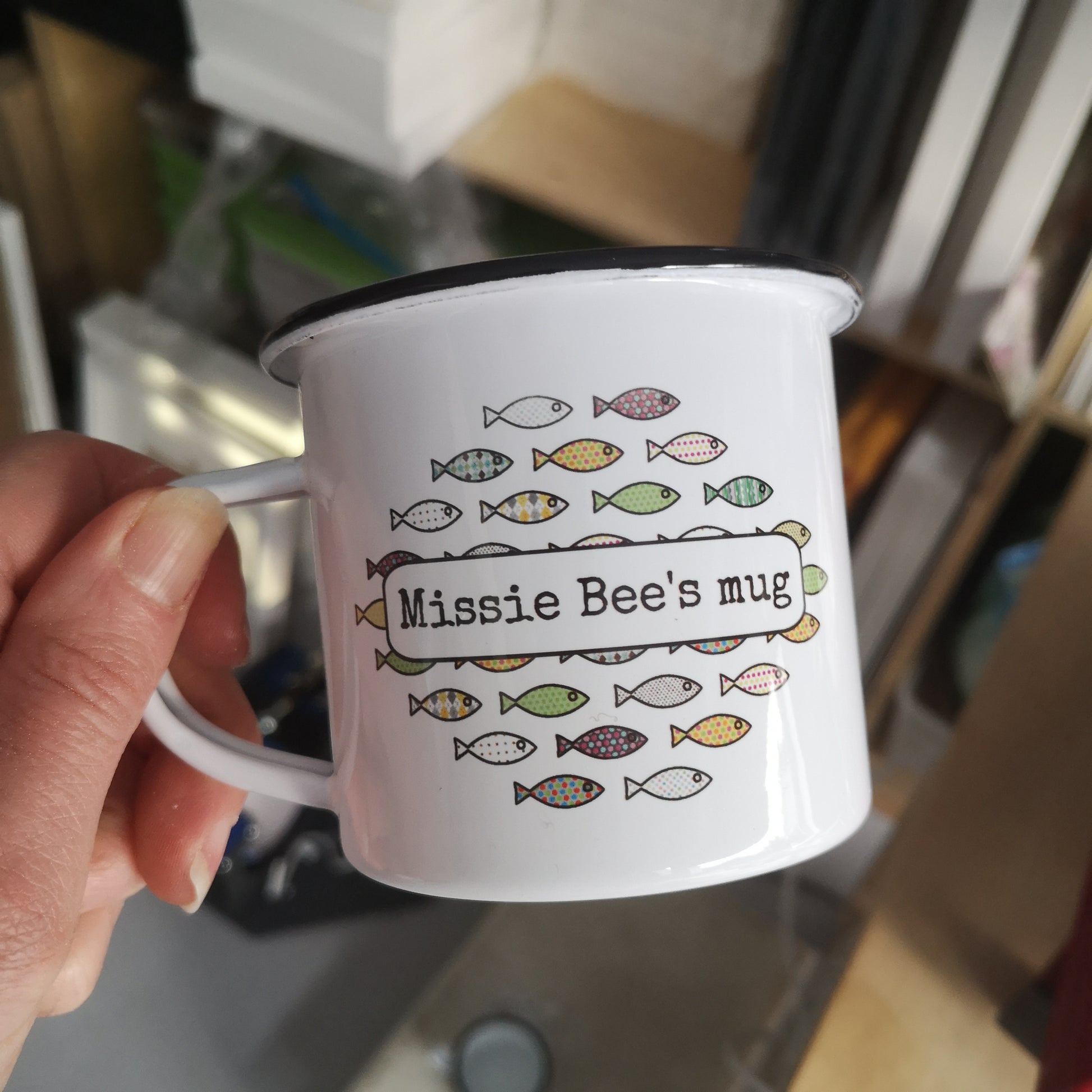 A different view of a white enamel mug, with a colourful shoal of fish on the front, overlayed with the owner's name.