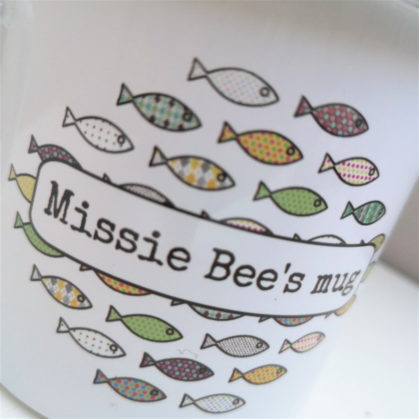 A close up photo of a white enamel mug, with a colourful shoal of fish on the front, overlayed with the owner's name.
