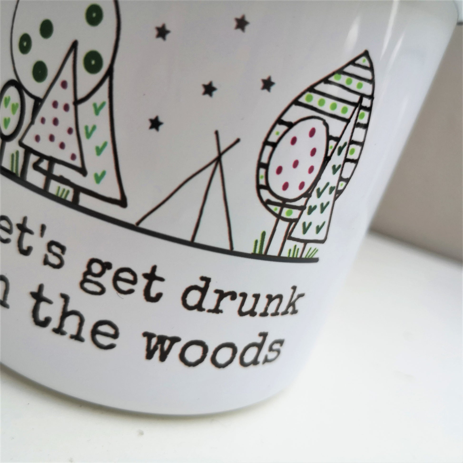 A close up of a white steel enamel mug with a black rim, with a handdrawn picture of a forest with a tent in the middle under stars, and written underneath in black type font - let's get drunk in the woods.