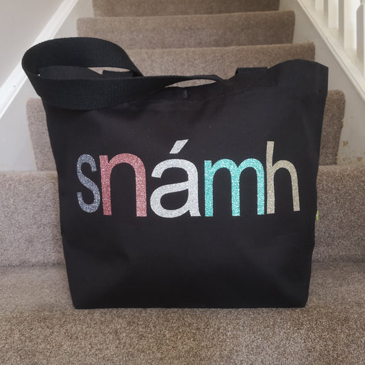  A black organic cotton tote with toggle closing with the following text on the front in various glitter colours in type font - SNÁMH