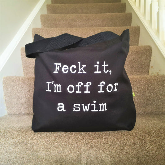 A black organic cotton tote with toggle closing with the following text on the front in white type font - FECK IT, I'M OFF FOR A SWIM