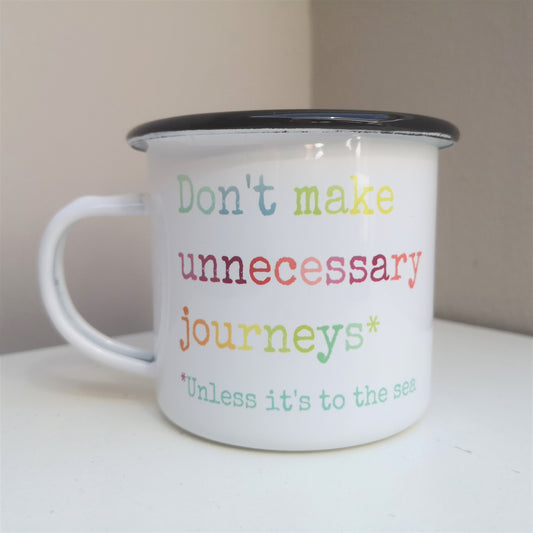A white steel enamel mug with the Teresa Mannion catchphrase DON'T MAKE UNNECESSARY JOURNEYS - UNLESS IT'S TO THE SEA written in rainbow coloured type font below