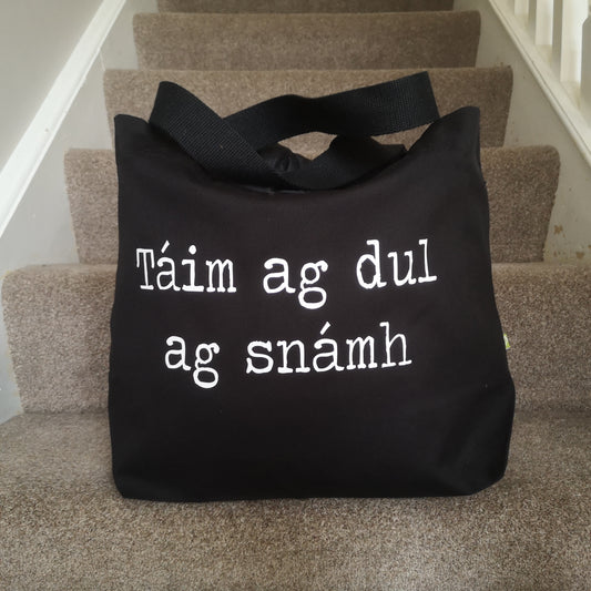 A black organic cotton tote with toggle closing with the following text on the front in white type font -"Táim ag dul as snamh"