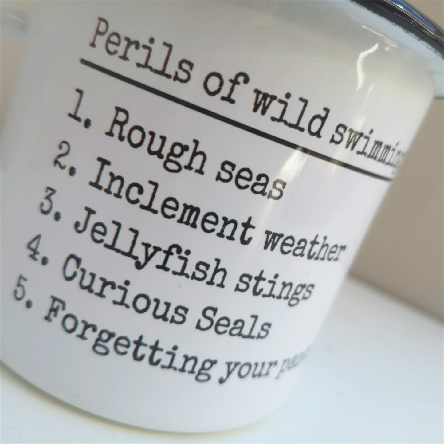 A close up photo of a White enamel mug with a black rim with the following on the front - Perils of wild swimming: Rough seas, Inclement weather, Jellyfish stings, curious seals, Forgetting your pants.