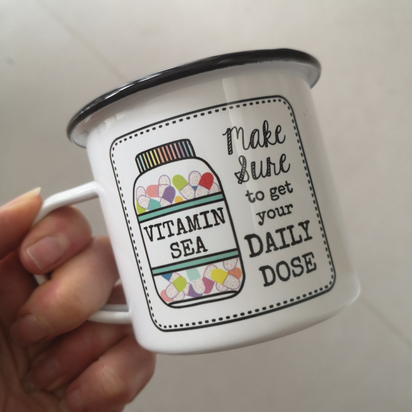 A photo of someone holding a White enamel mug with a black rim with the following on the front - a framed hand drawn image of a vitamin jar filled with brightly coloured pills and text to the right of it that says "make Sure to get your daily dose". The bottle has a VITAMIN SEA label on it.