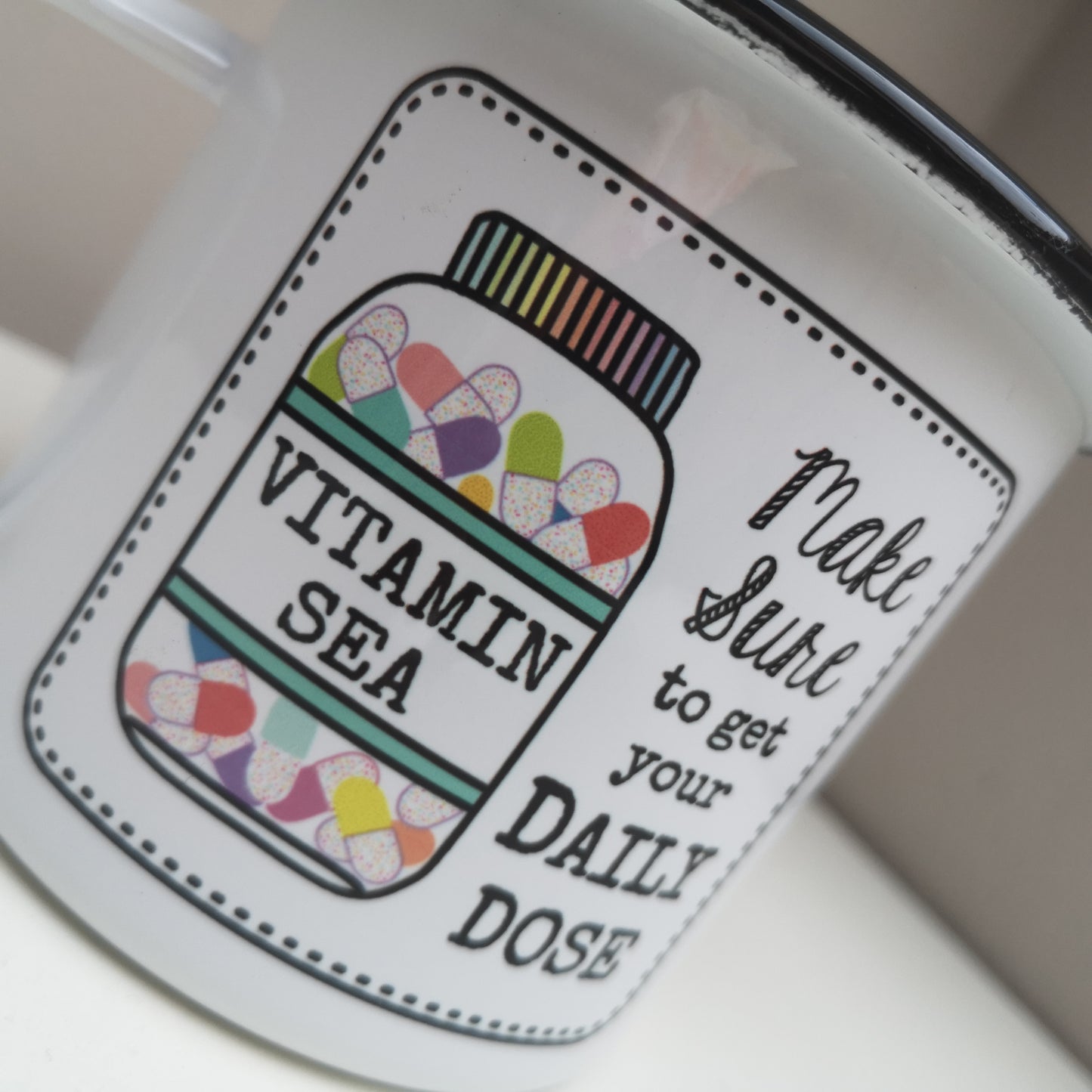 A close up photo of a White enamel mug with a black rim with the following on the front - a framed hand drawn image of a vitamin jar filled with brightly coloured pills and text to the right of it that says "make Sure to get your daily dose". The bottle has a VITAMIN SEA label on it.