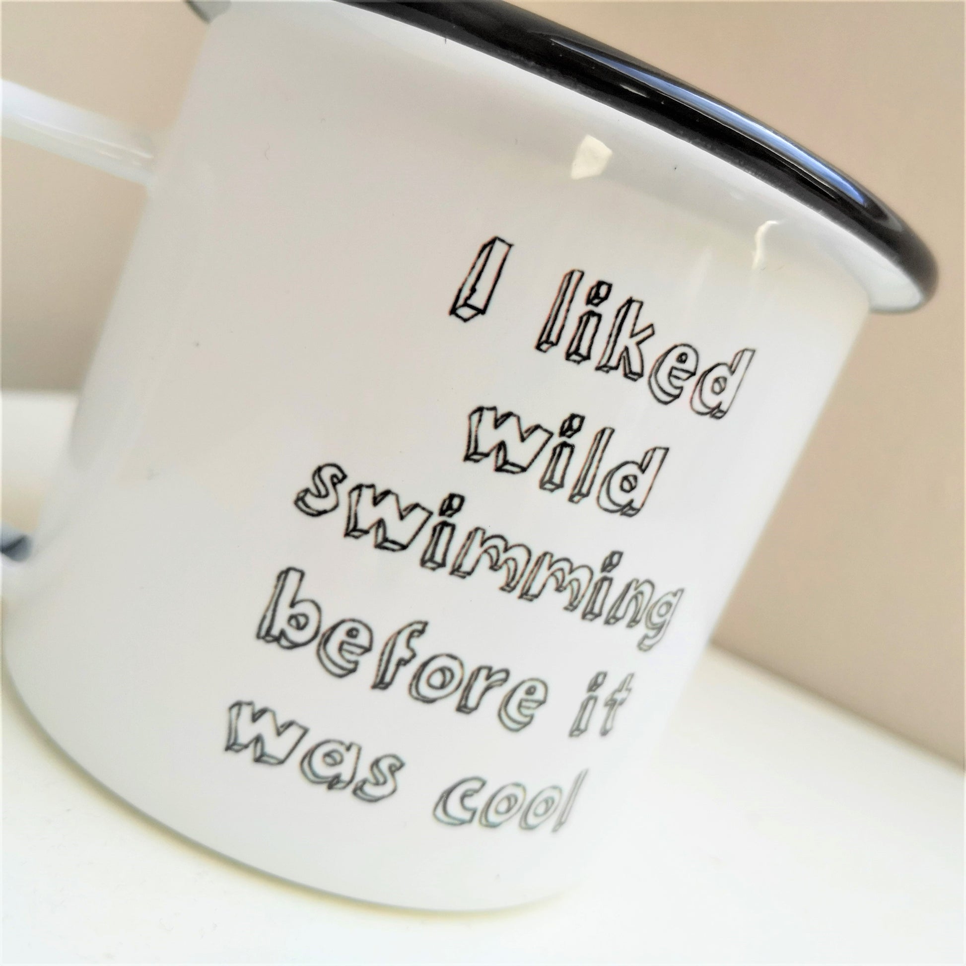 A close up of a white steel enamel mug with a black rim, with the following text in black 3d writing - I LIKED WILD SWIMMING BEFORE IT WAS COOL