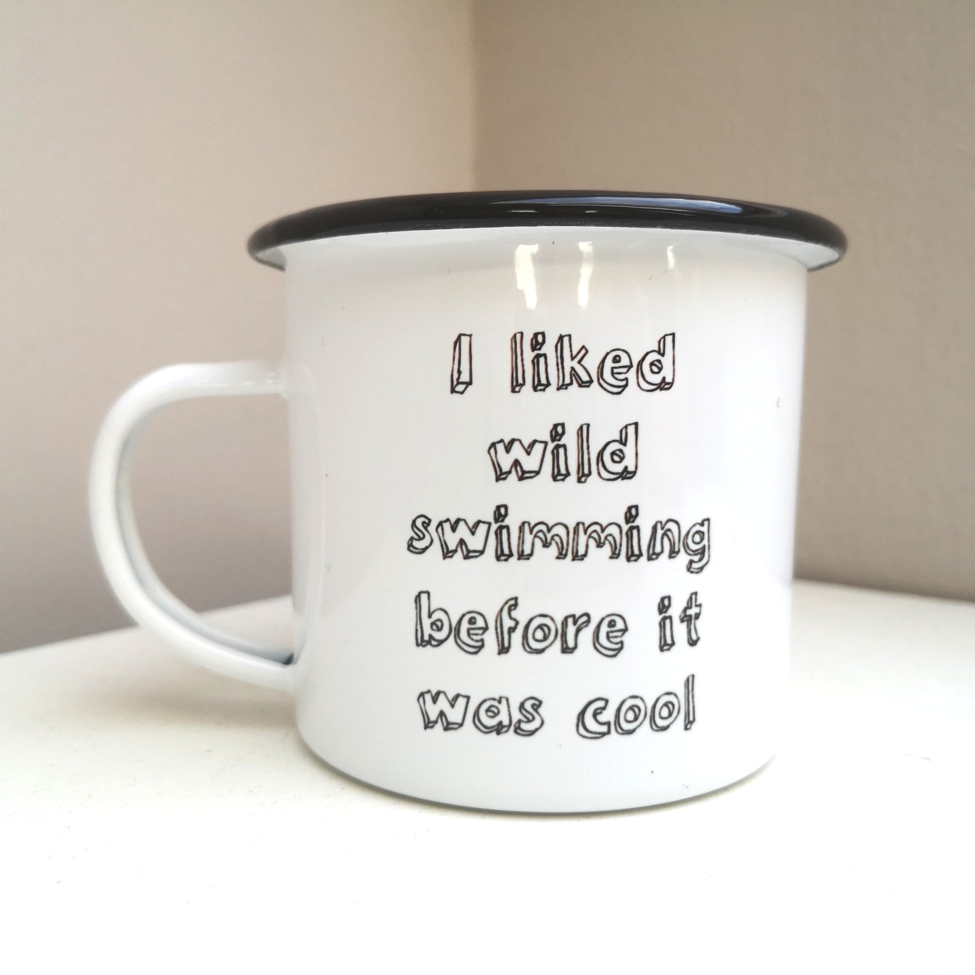 A white steel enamel mug with a black rim, with the following text in black 3d writing - I LIKED WILD SWIMMING BEFORE IT WAS COOL