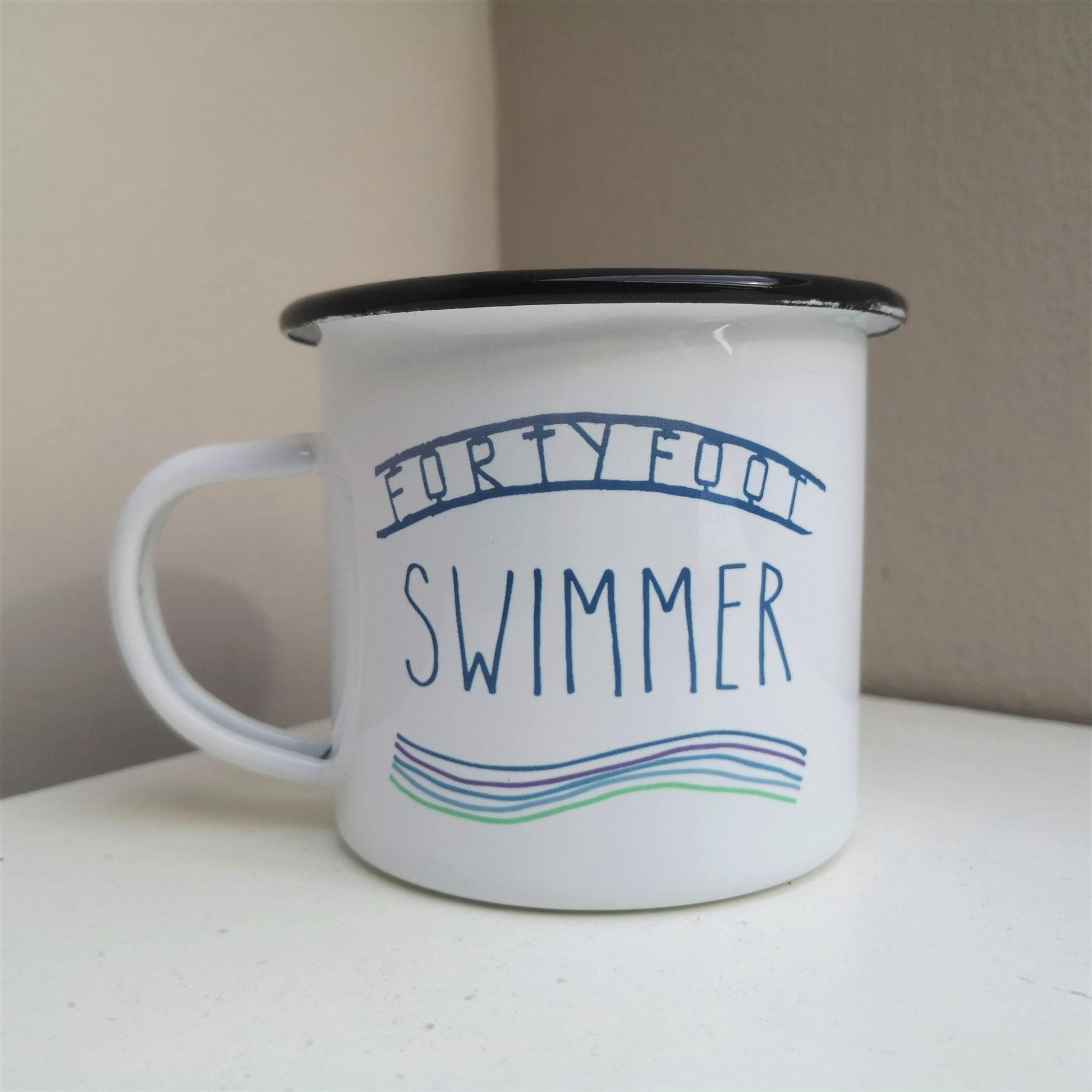 A white steel enamel mug with black rim, with FORTY FOOT SWIMMER written on it and some waves below, all in aquamarine tones.  The FORTY FOOT is the iconic sign that you see down at the bathing place