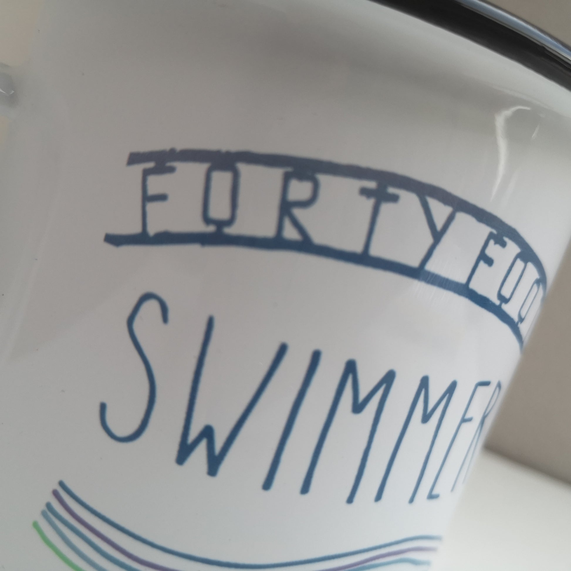 A close up of a white steel enamel mug with black rim, with FORTY FOOT SWIMMER written on it and some waves below, all in aquamarine tones.  The FORTY FOOT is the iconic sign that you see down at the bathing place