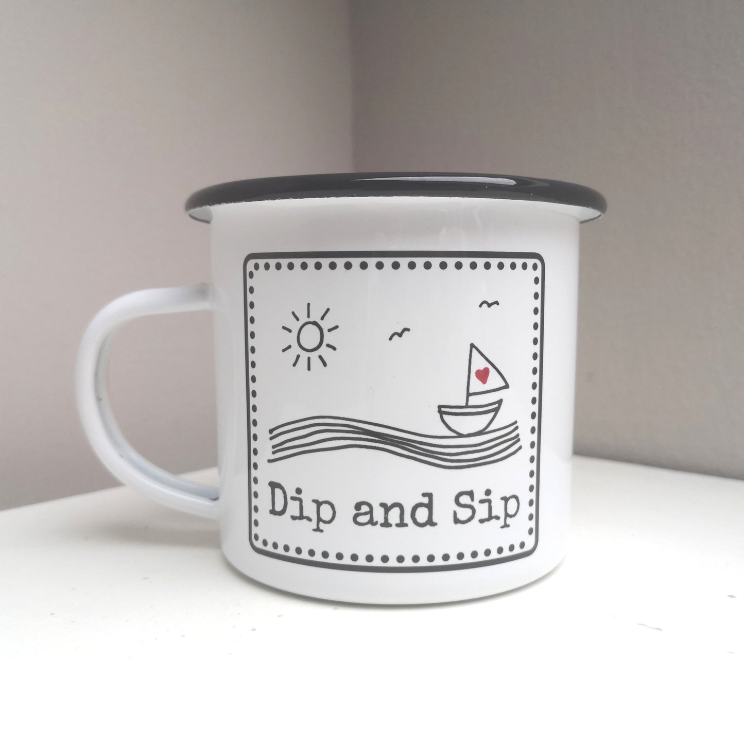 A white steel enamel mug with a year round swimmer's mantra on it - dip and sip.  Photo shows the black and white design with a sailboat