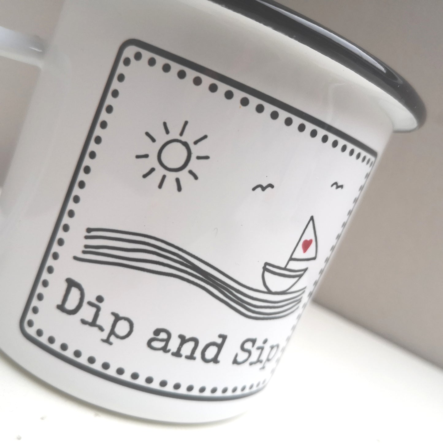 A white steel enamel mug with a year round swimmer's mantra on it - dip and sip. Photo shows a close up of the black and white design with a sailboat