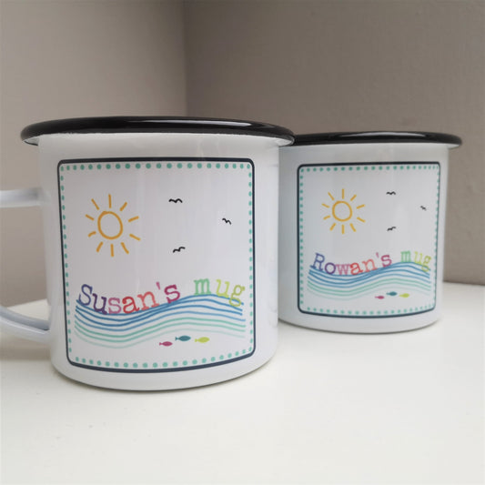 A photo showing two white enamel mugs featuring a colourful framed seaside scene, with the mug owner's name riding on the waves
