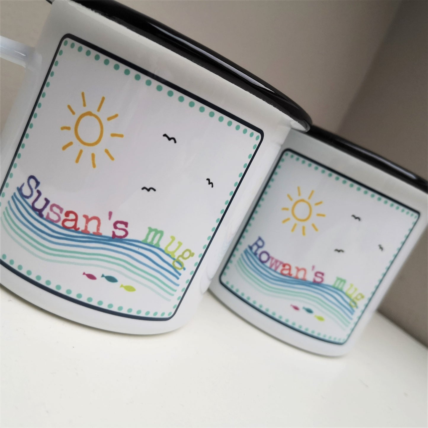 A close up photo of 2 white enamel mugs featuring a colourful framed seaside scene, with the mug owner's name riding on the waves!