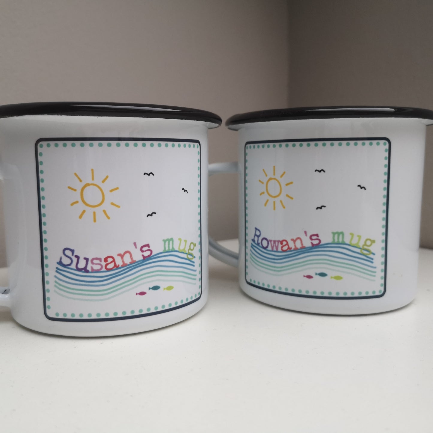A close up view of 2 white enamel mugs, featuring a colourful framed seaside scene, with the mug owner's name riding on the waves!