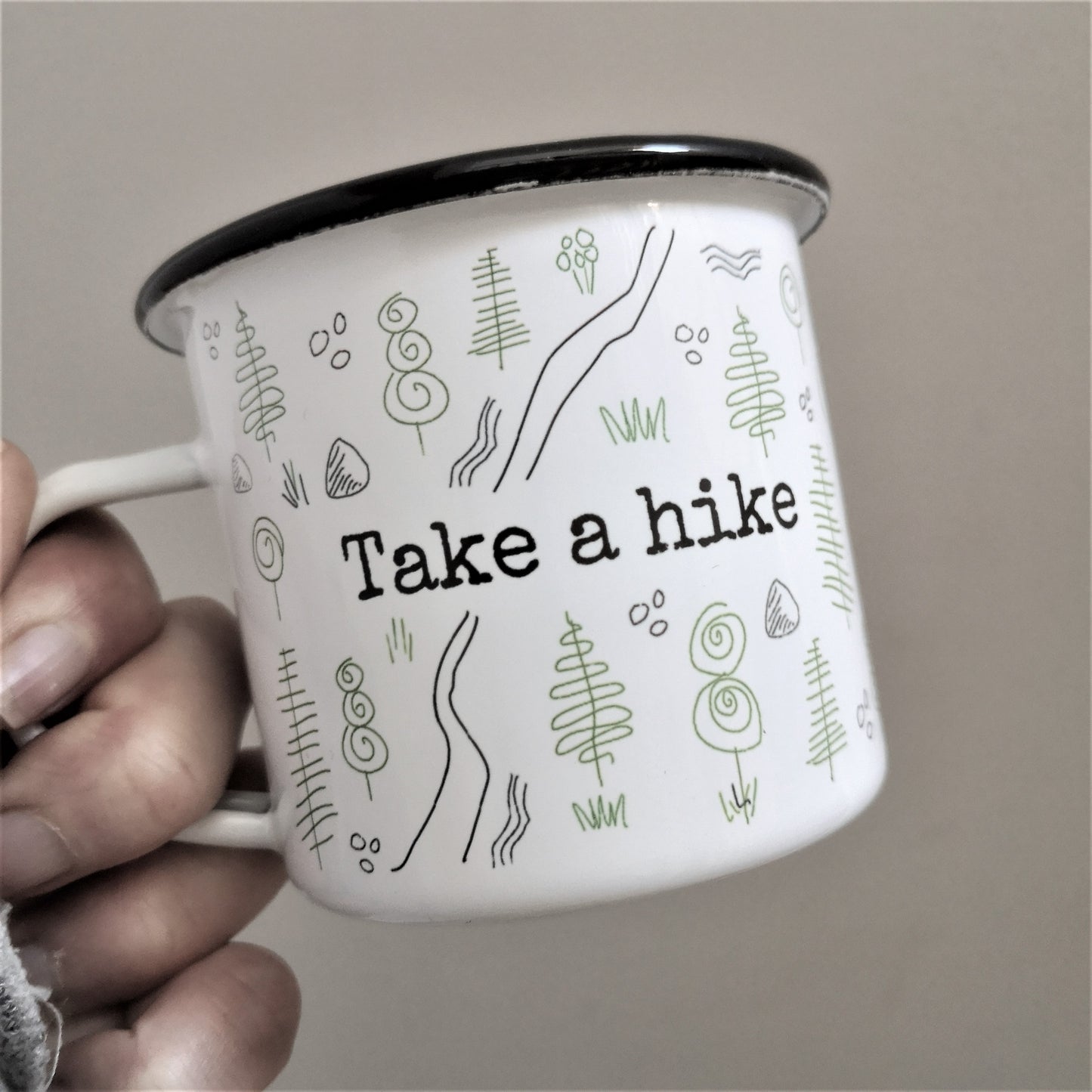 A White enamel mug with a black rim with the following design wrapping around the outside of the mug - a forest scene with a rough path through it and TAKE A HIKE written in the centre on both the back and front of the mug.
