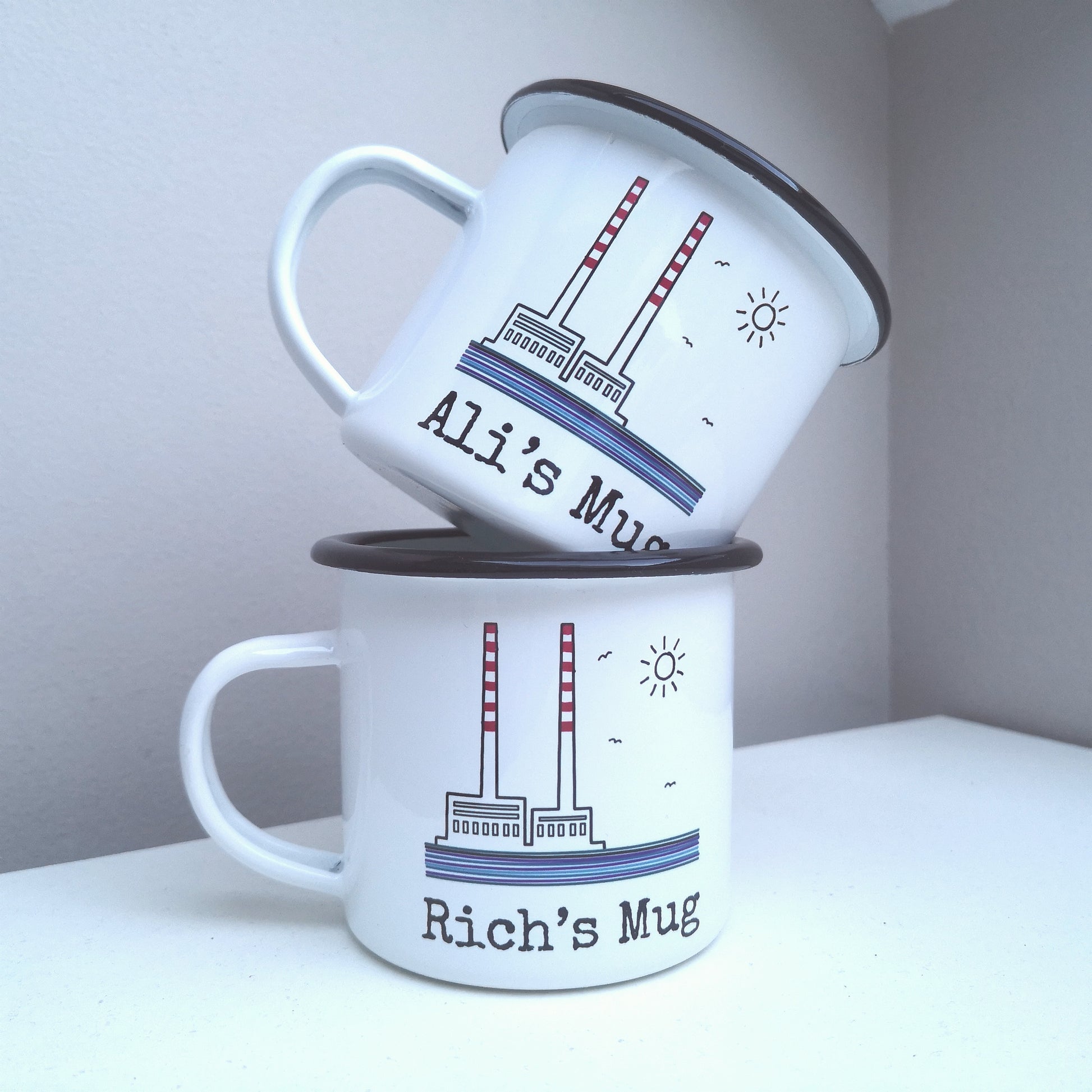 A photo showing 2 personalised white steel enamel mug with a hand drawn picture of the Poolbeg towers on it. The mugs are stacked on top of each other.