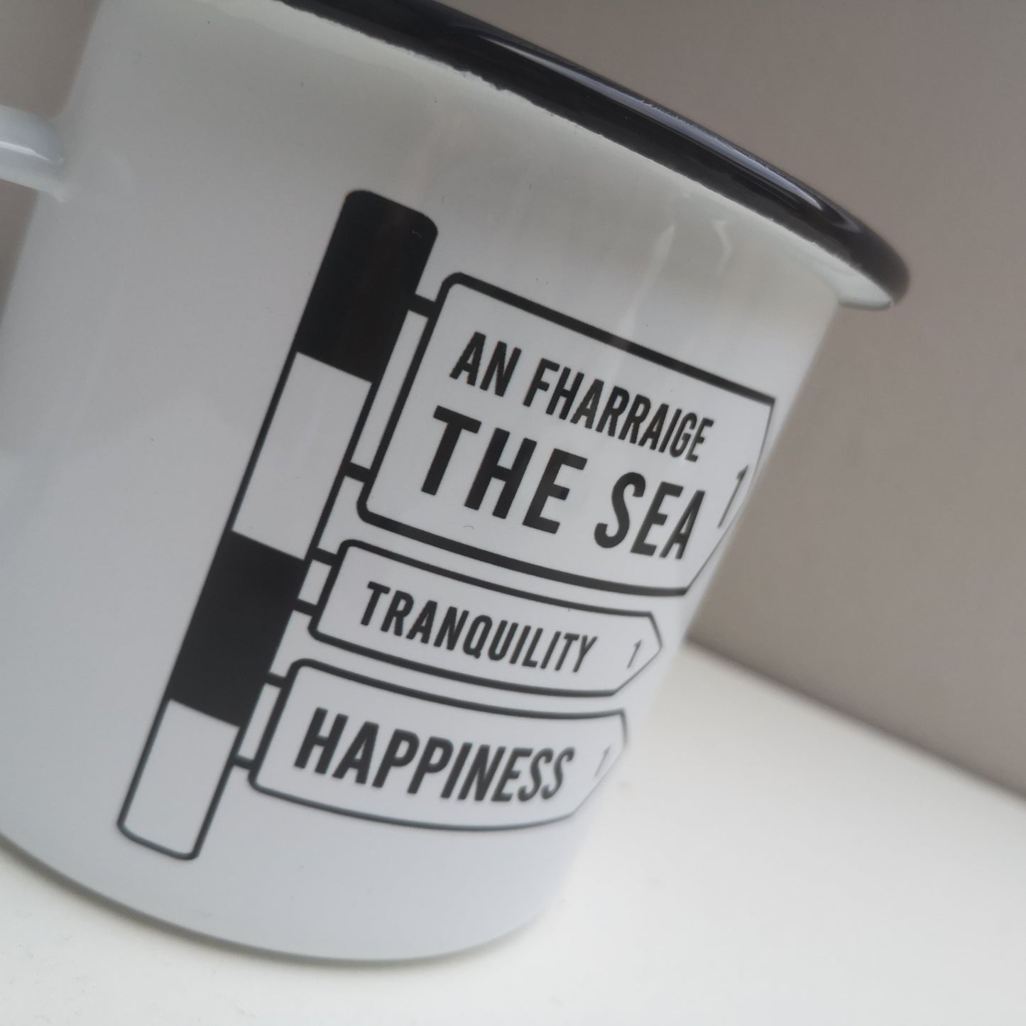 A close up of a White enamel mug with a black rim with the following on the front -an old Irish Roadsign pointing to the right with arrows for The Sea, Tranquility and Happiness