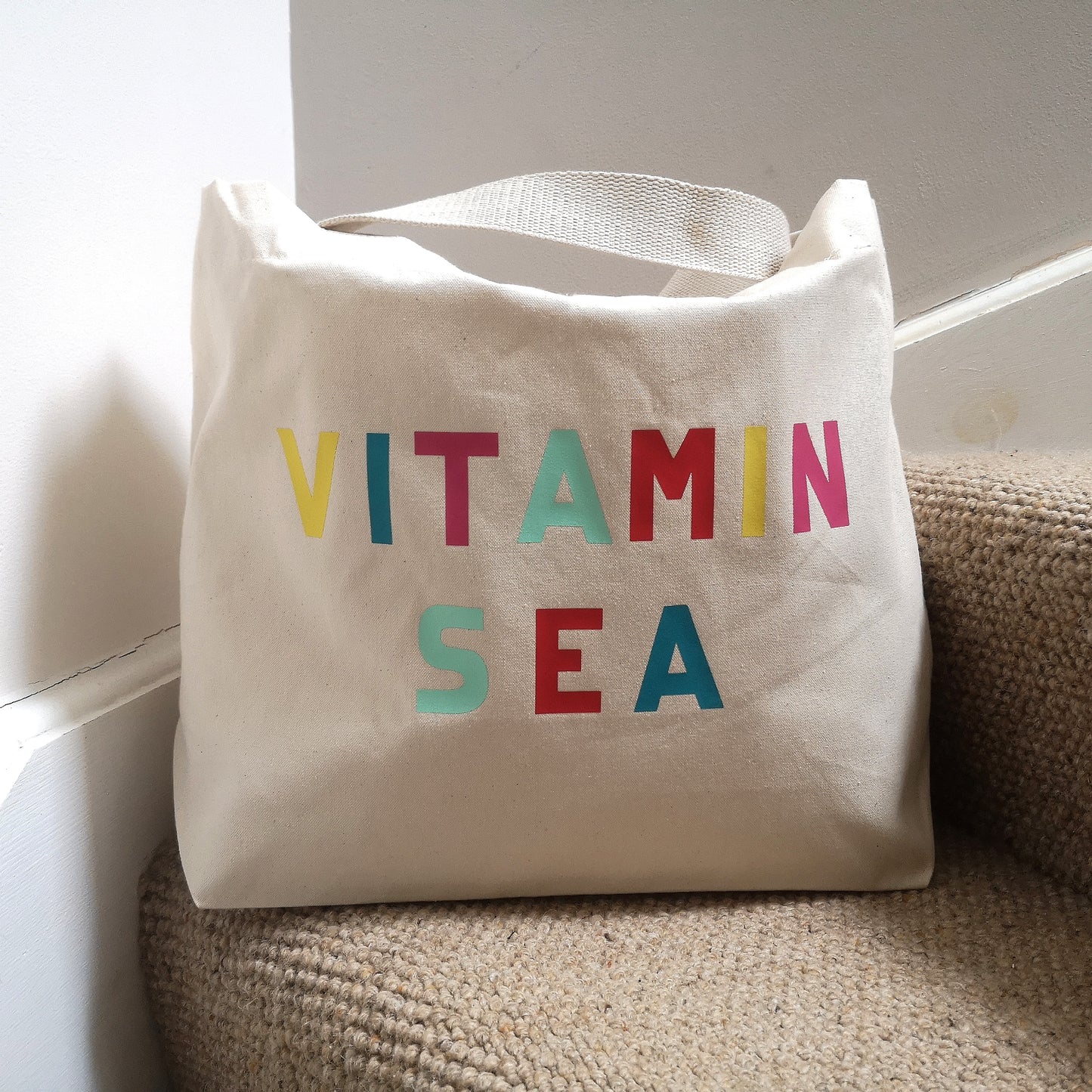 A natural cotton organic cotton tote with toggle closing with VITAMIN SEA in different coloured bright lettering on the front