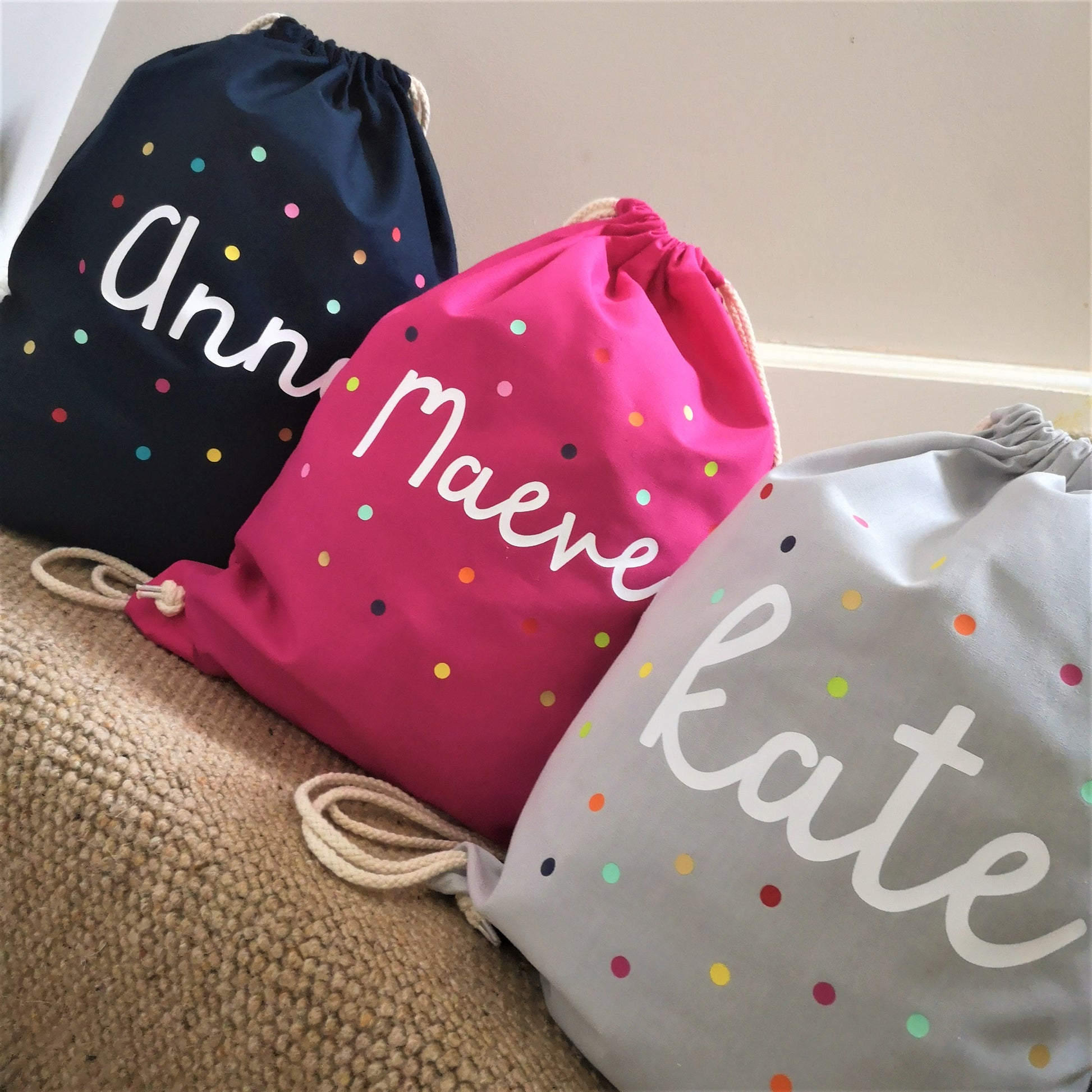 A photo showing the 3 bag colour choices of a personalised light cotton drawstring bag with dots and personalised with a name.  The 3 colour choices are french navy, fuschia pink and light grey