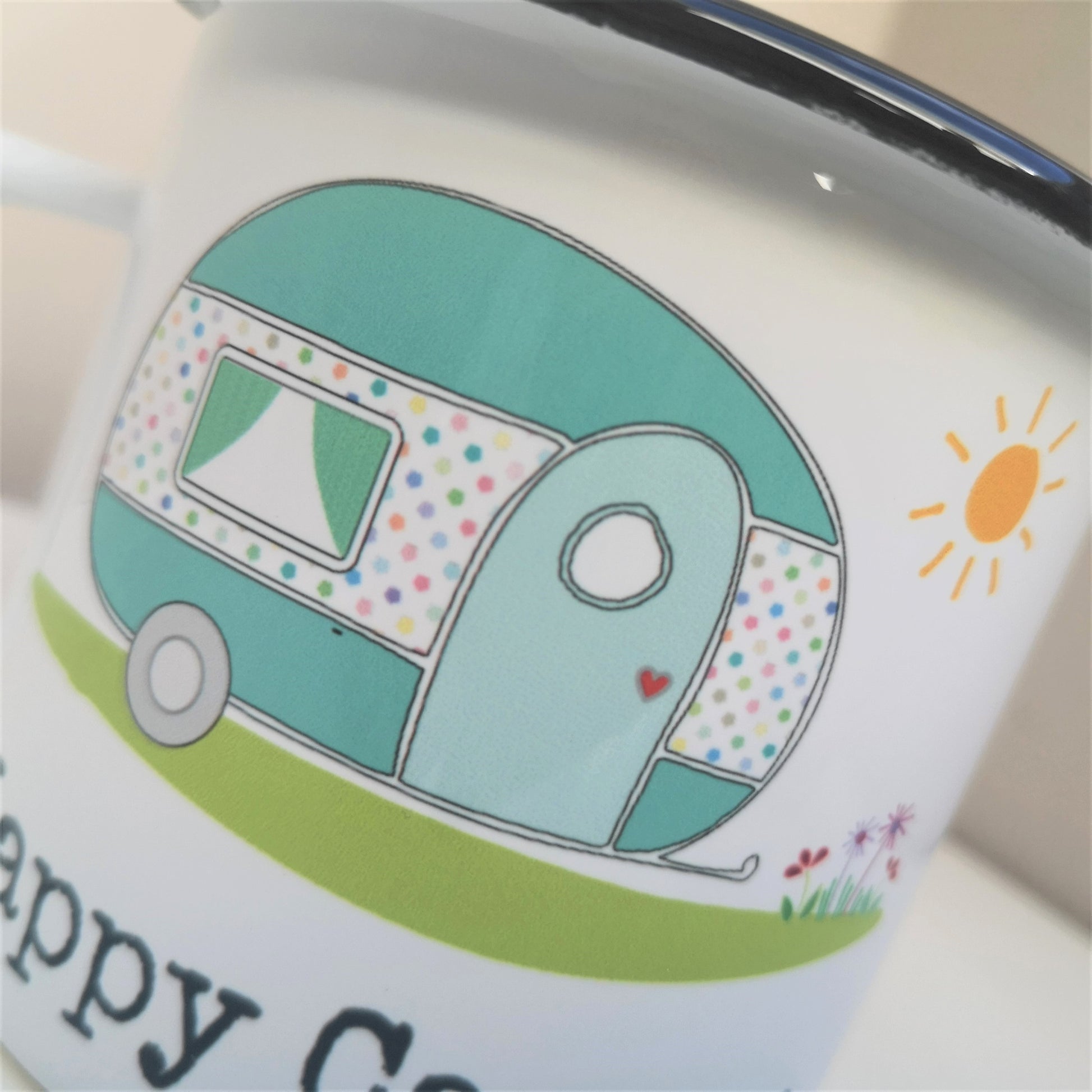 A close up of a white steel enamel mug with a cartoon picture of a caravan in pretty pastel colours, and below it written in type font is HAPPY CAMPER