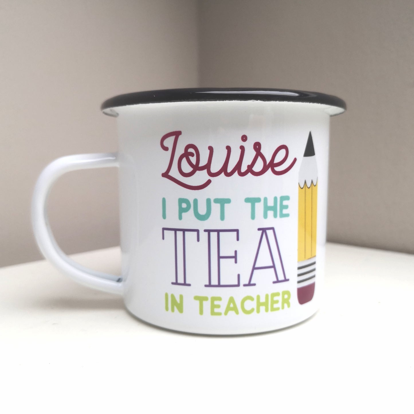 A White enamel mug with a black rim with the following on the front - <NAME> I out the tea in teacher and a big pencil to the right of the text