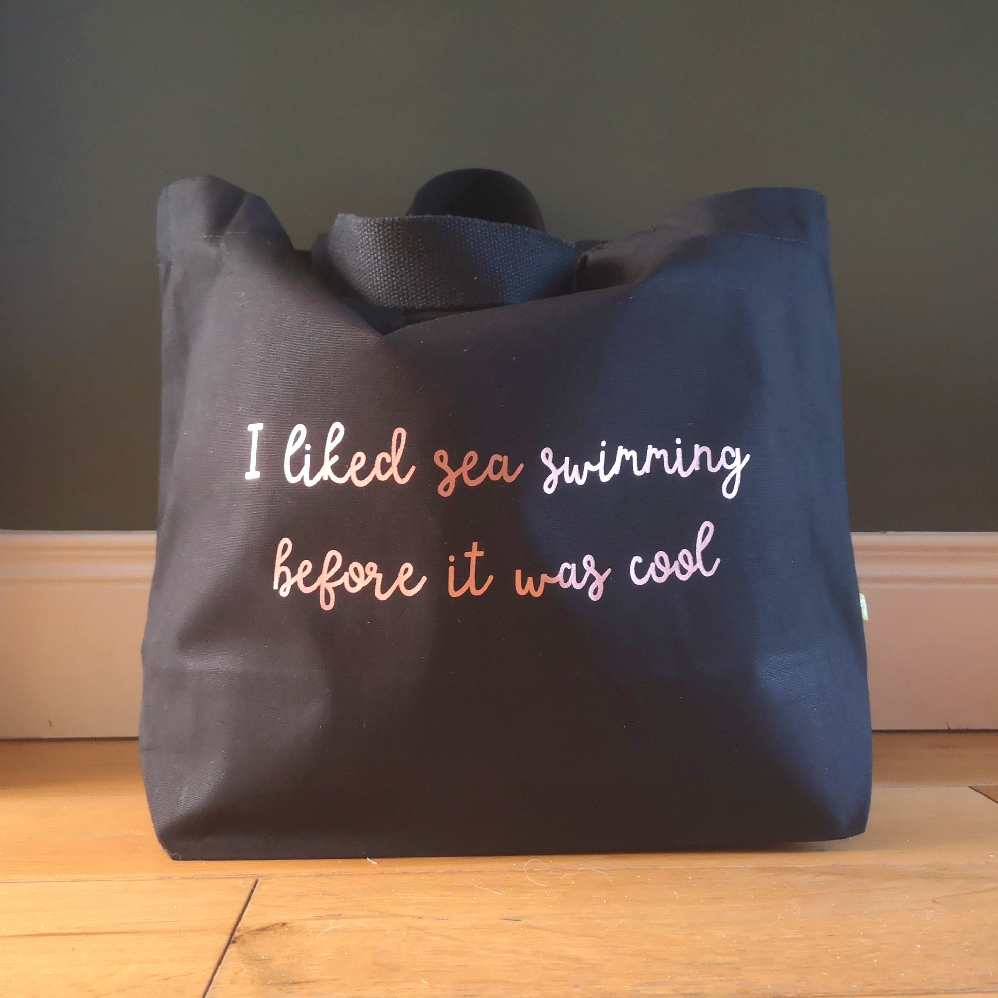 A black organic cotton tote with toggle closing with the following text on the front in rose gold handwritten font - I liked sea swimming before it was cool 